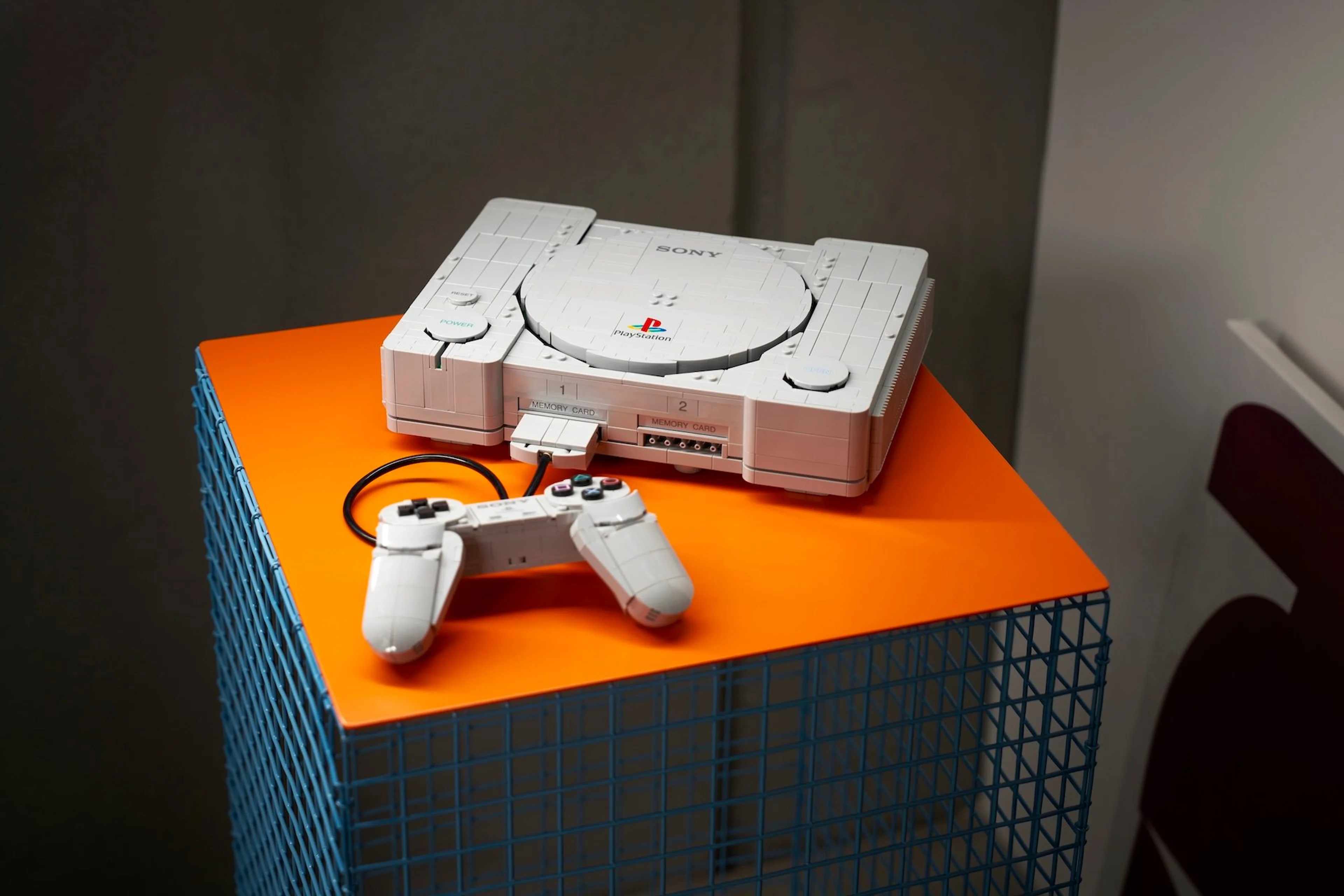 The completed LEGO PlayStation set sits on an orange display stand with its controller in front.