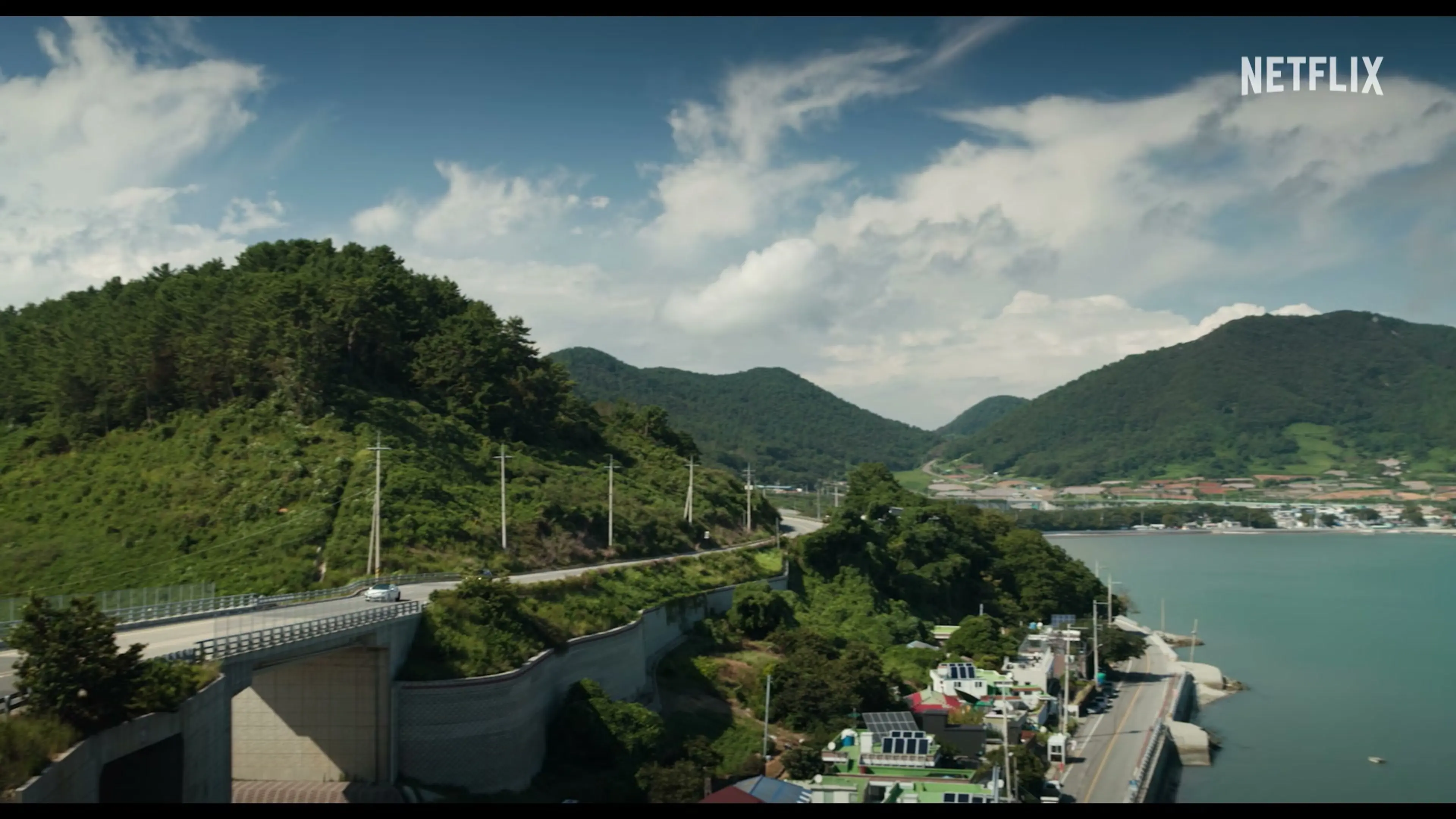 A road passes wooded hills beside a harbor with fishing boats.