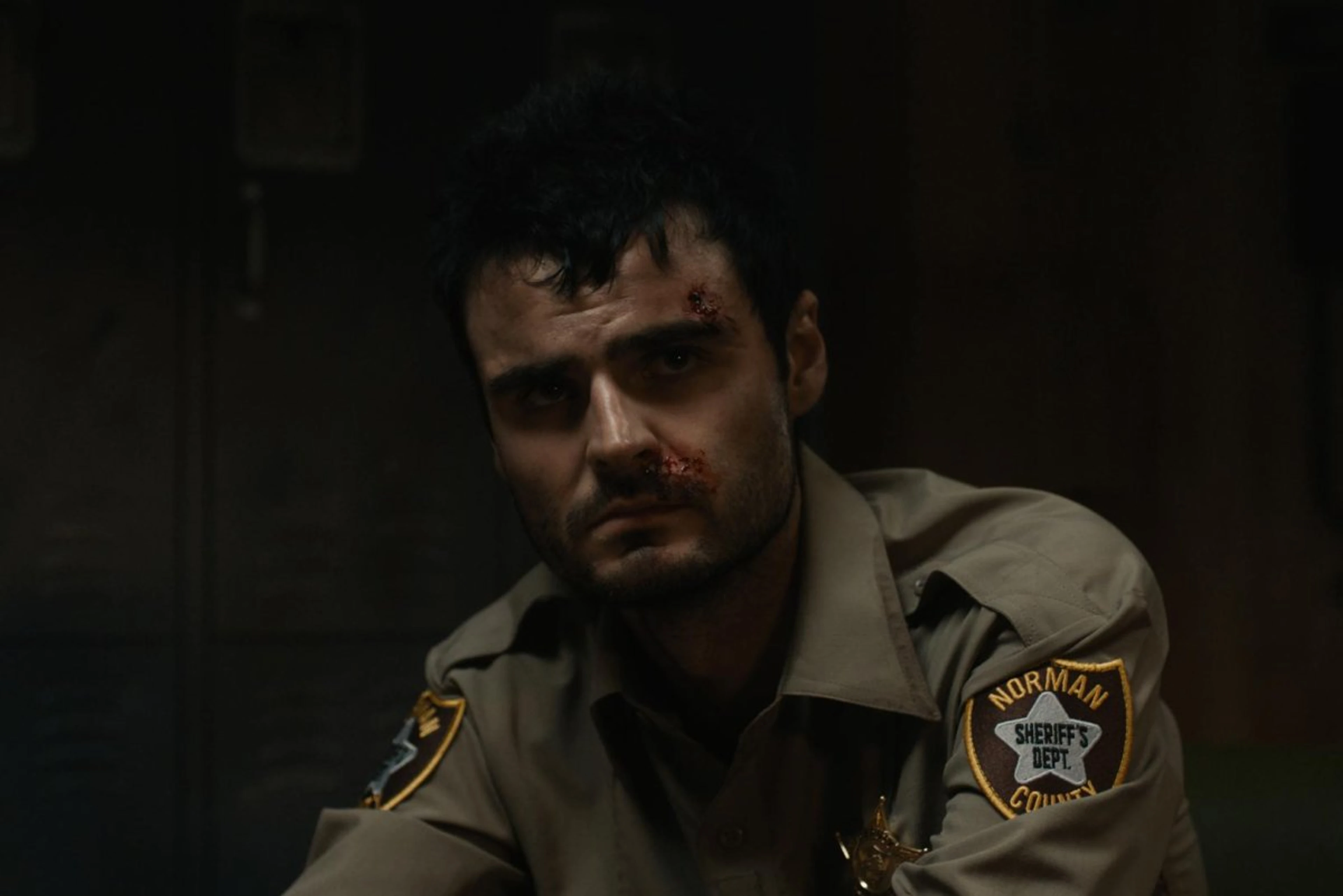 A bruised Duke Nicholson wears a sheriff’s uniform in a dark room.