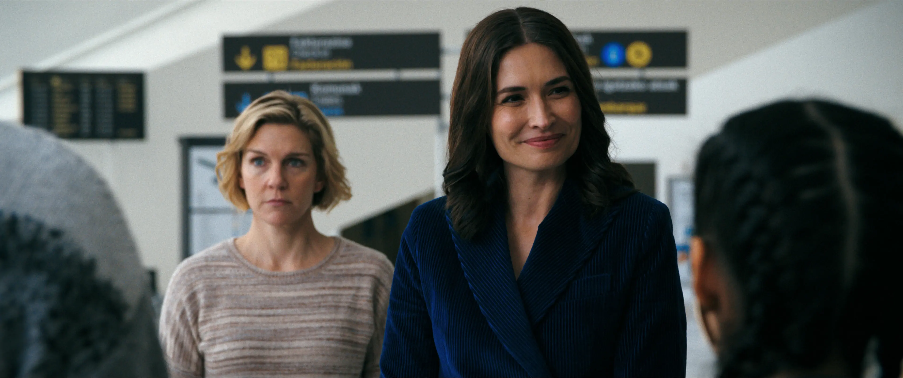 Rhea Seehorn stands behind Karolina Wydra in an airport scene from Pluribus.