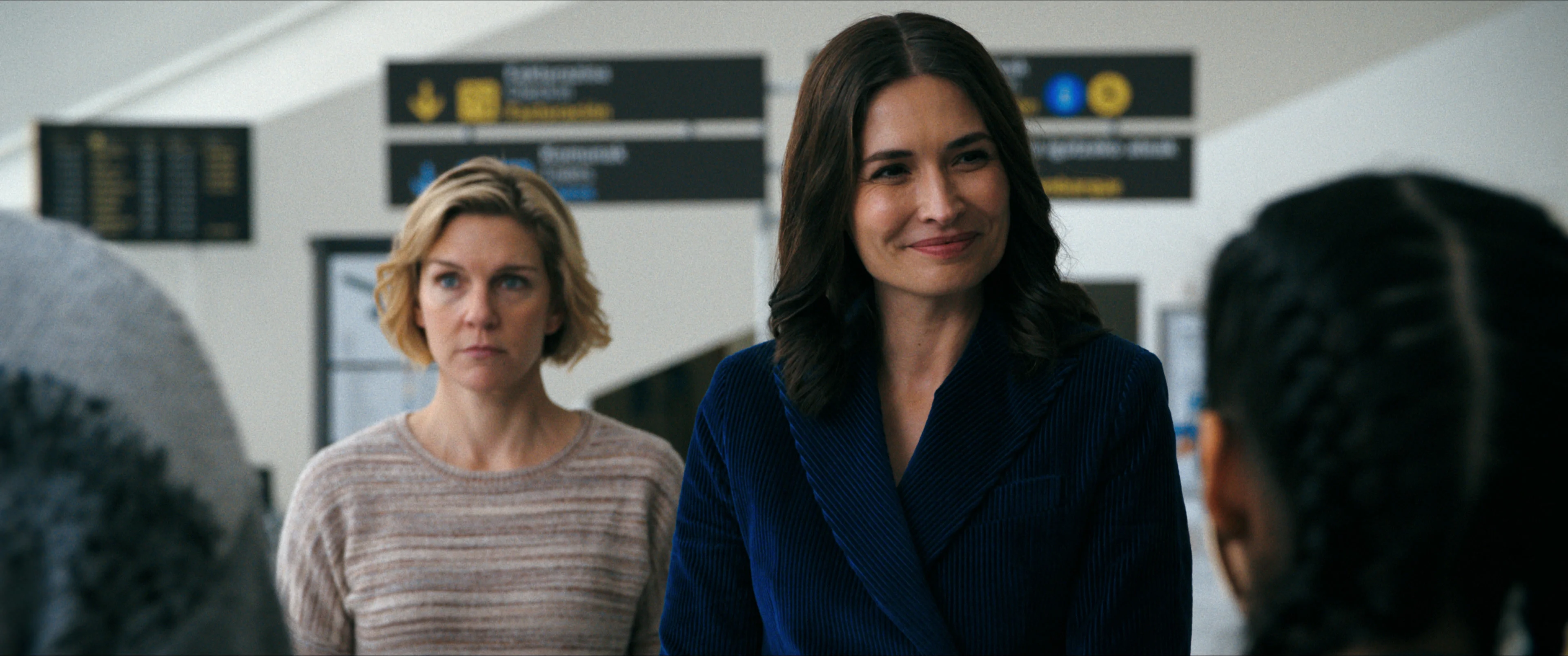 Rhea Seehorn stands behind Karolina Wydra in an airport scene from Pluribus.