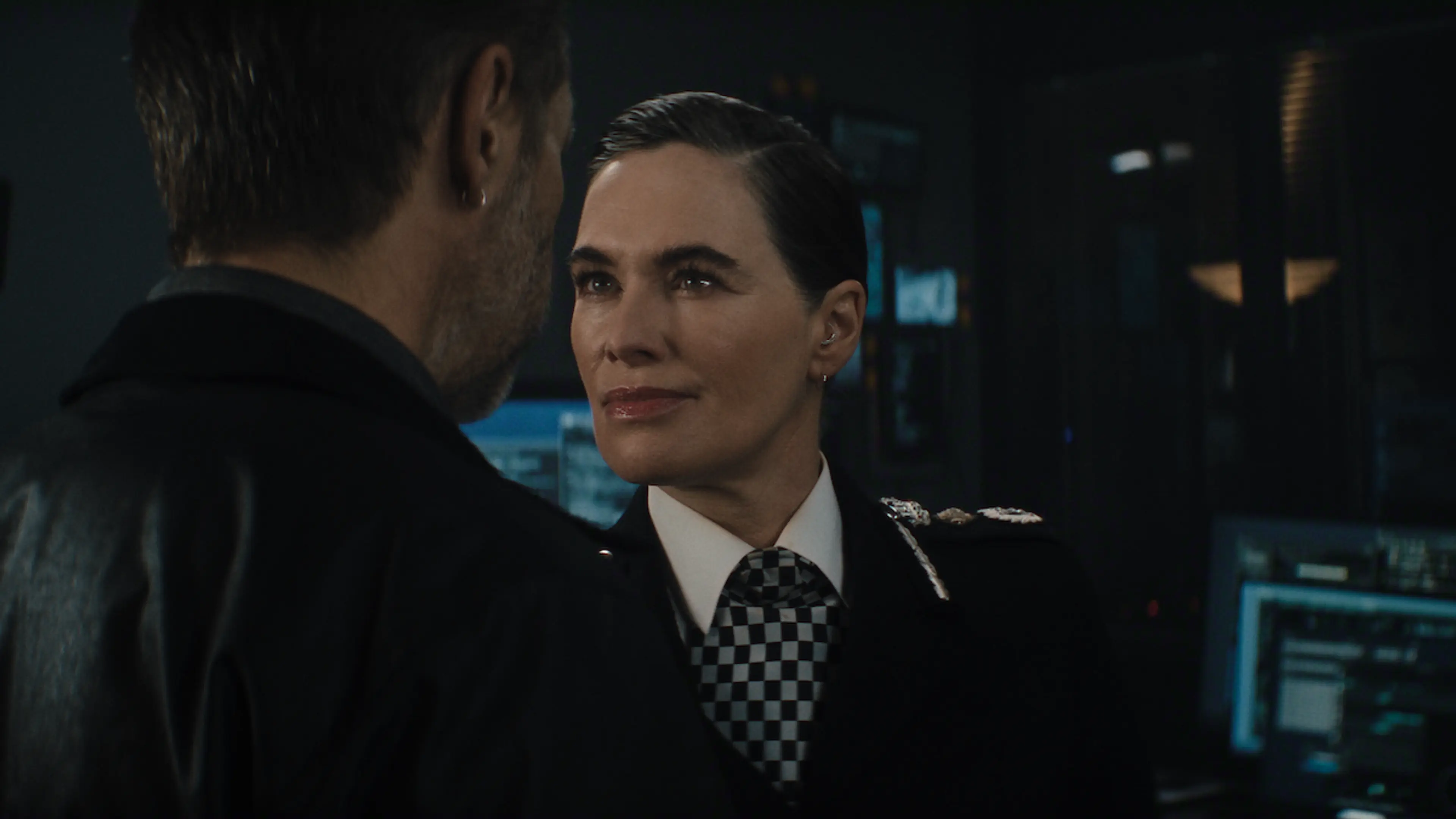 A senior police officer faces a colleague in a dark control room.
