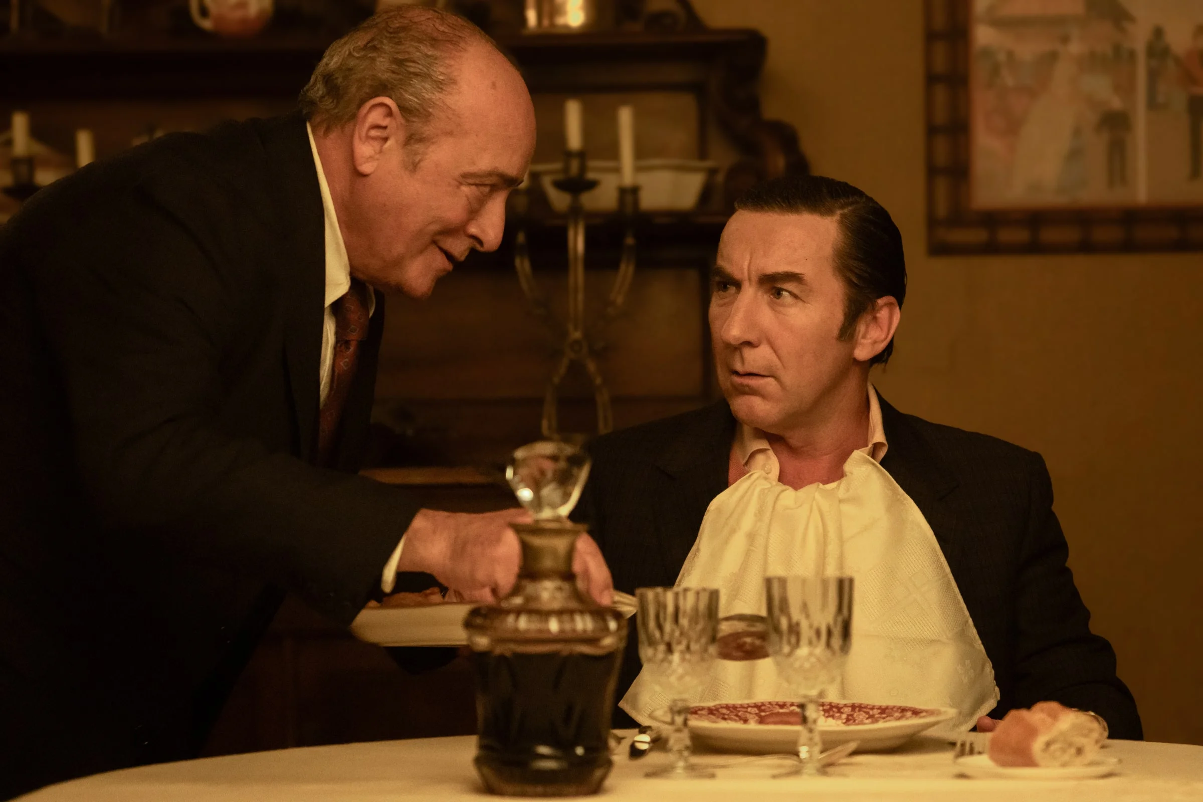 Don Leandro looks up as an older man talks to him across a dinner table.