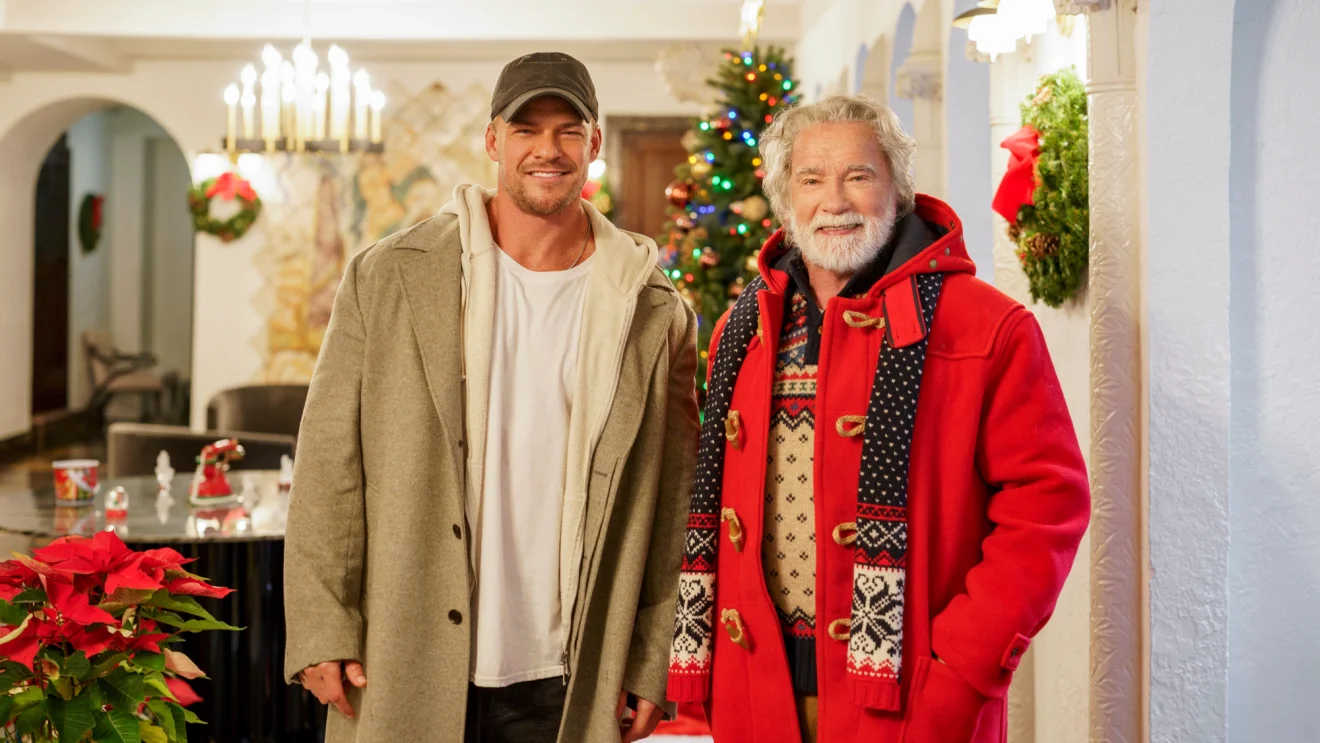 Alan Ritchson and Arnold Schwarzenegger stand together in a Christmas-decorated house.