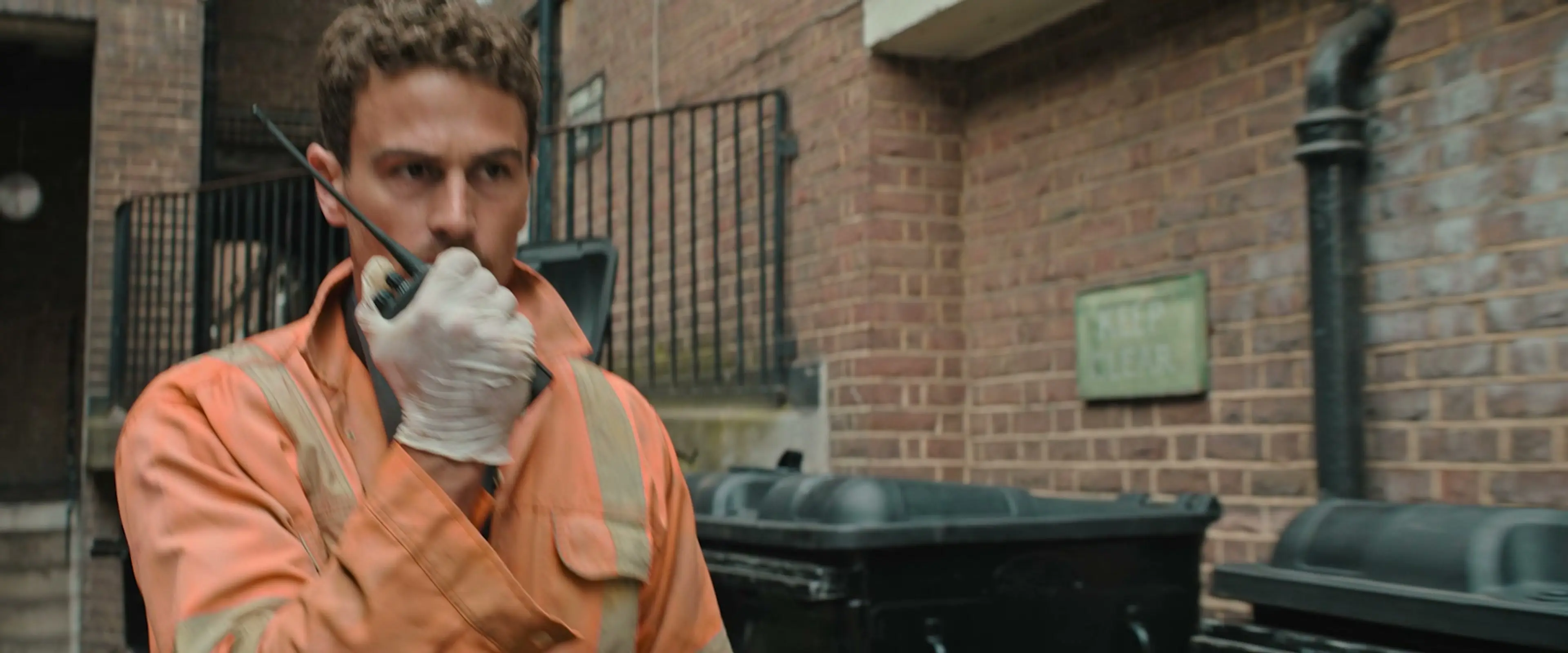 Karalis wears orange work clothes and speaks into a handheld radio outside a brick building.