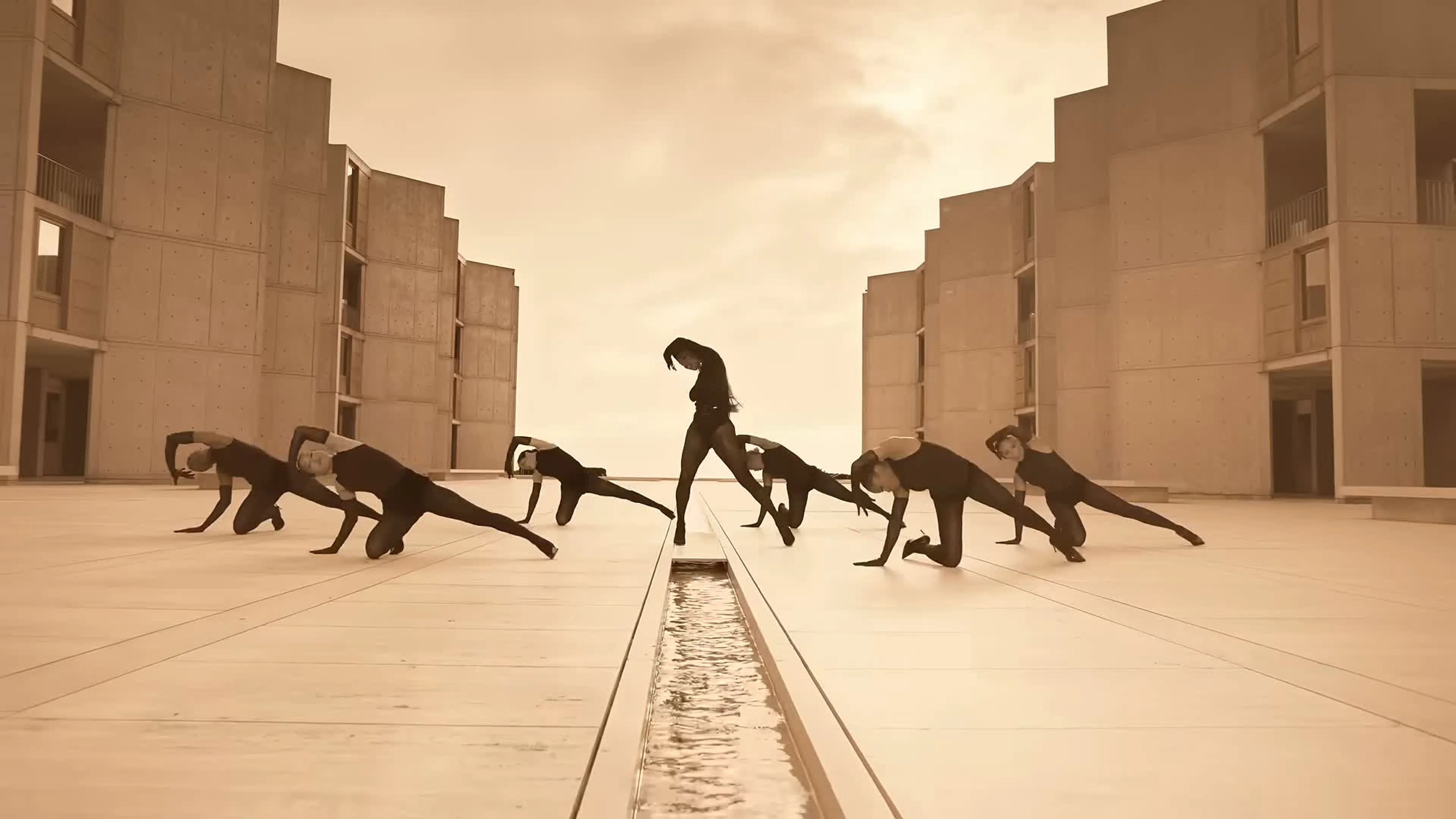 Victoria Monét and six dancers pose beside a channel of water outside a concrete building in Reach Out.