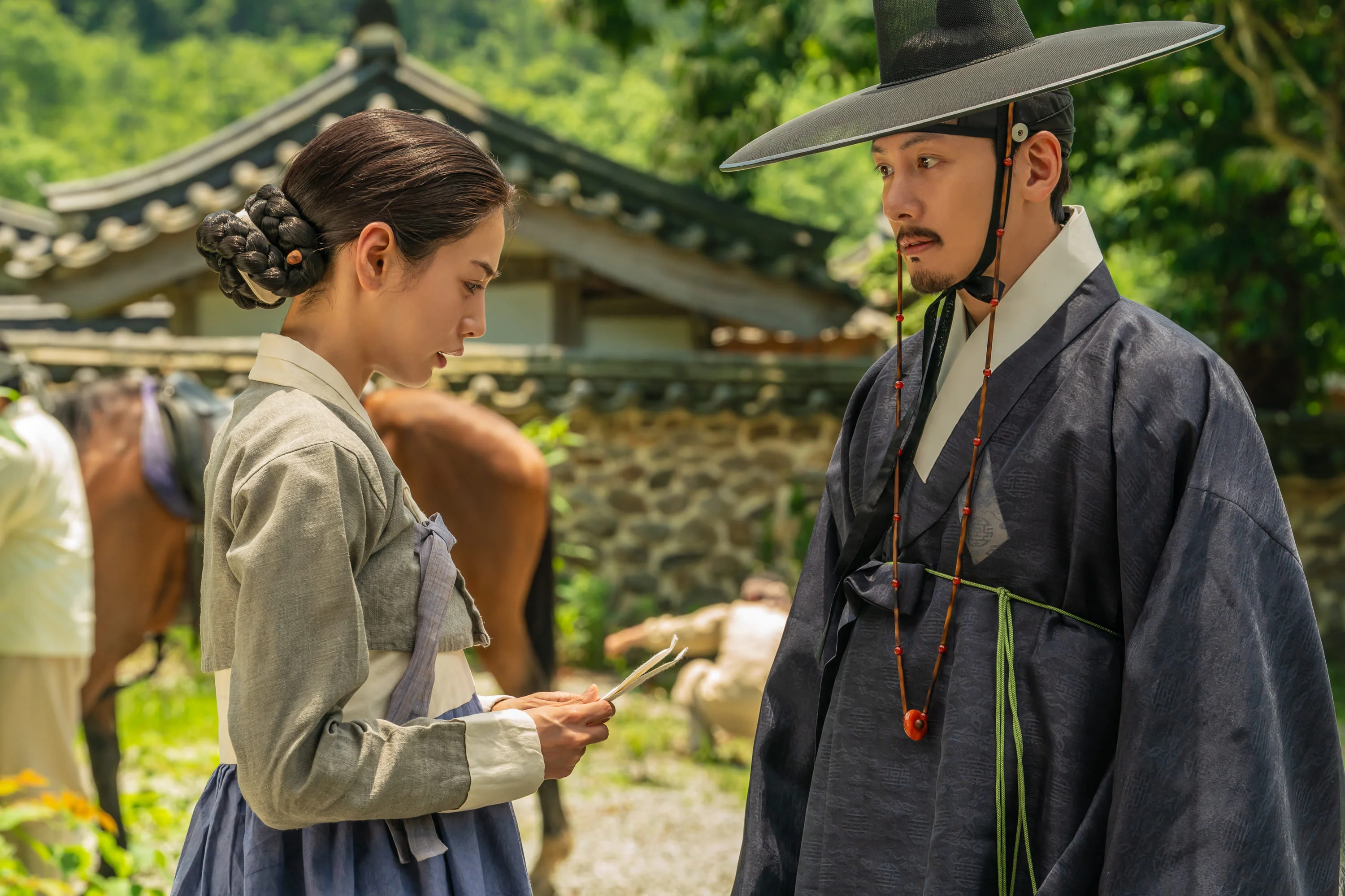 Hui-yeon holds a paper while Cho Won watches her in a courtyard.