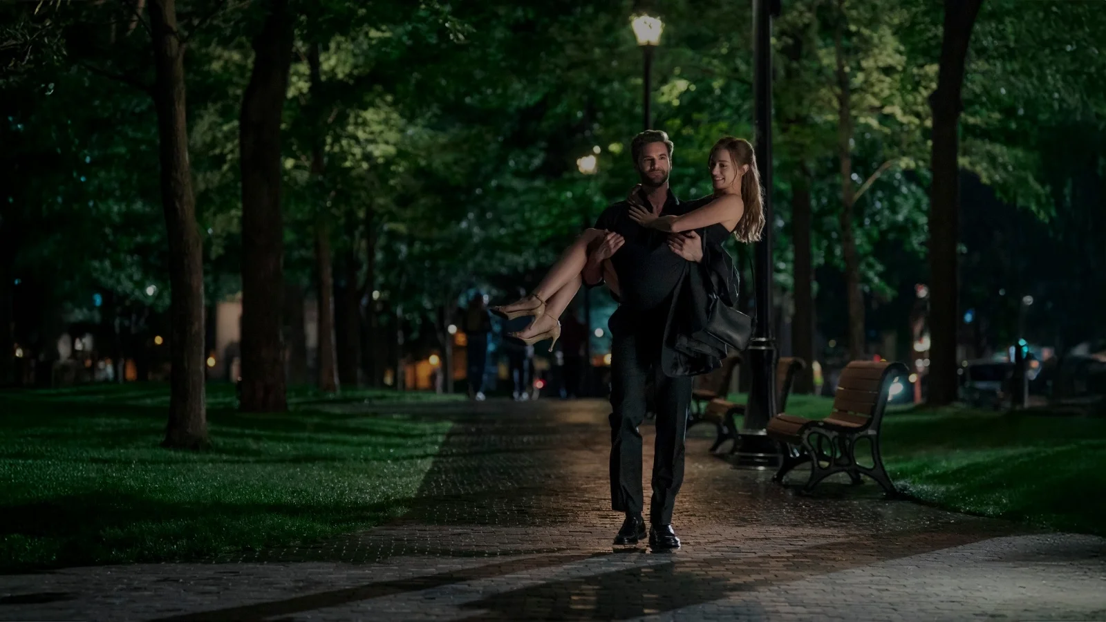 Tom Bateman carries Lili Reinhart along a park path in The Love Hypothesis.