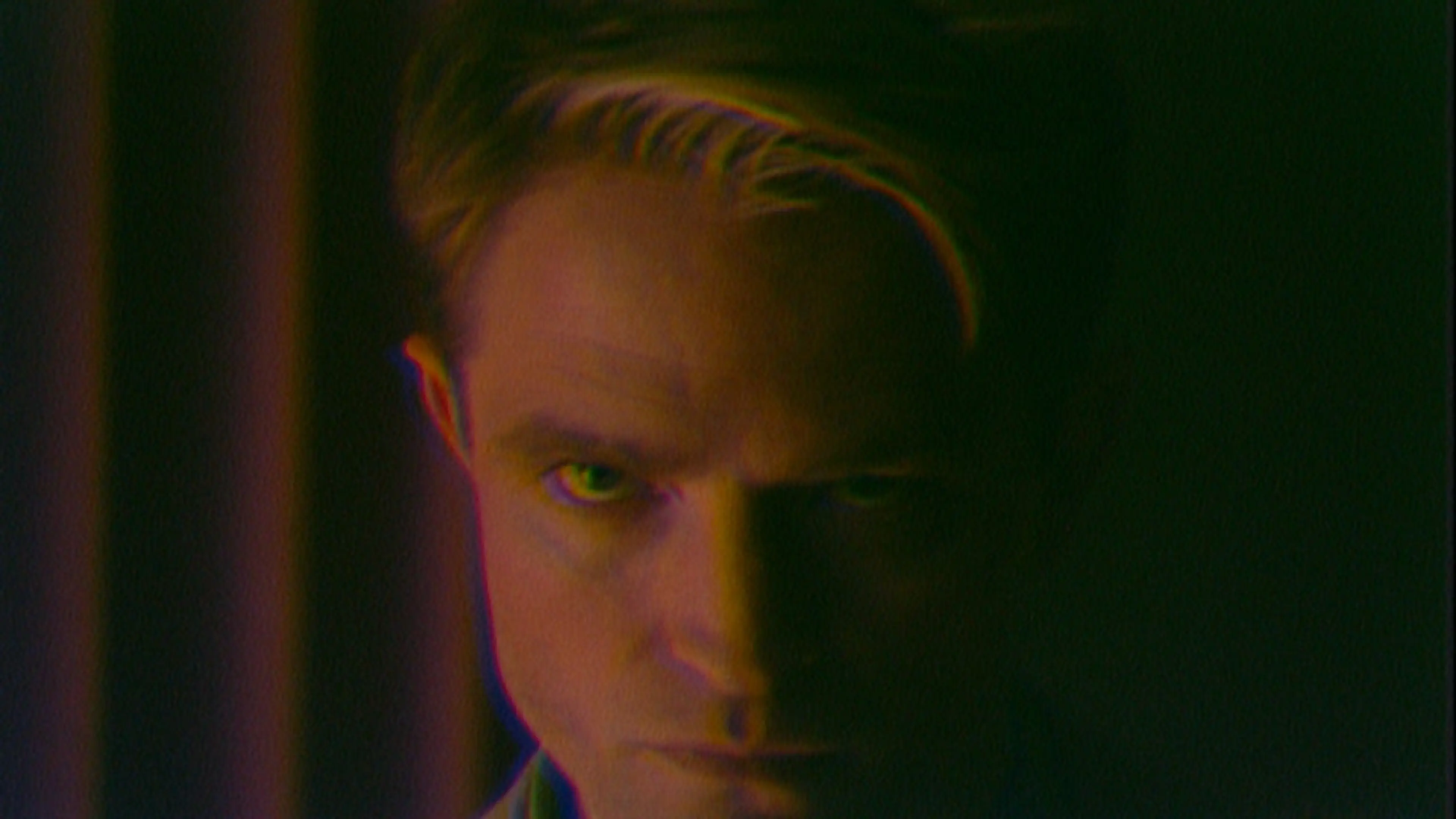 A dark close-up of Pattinson’s face under purple and green light.