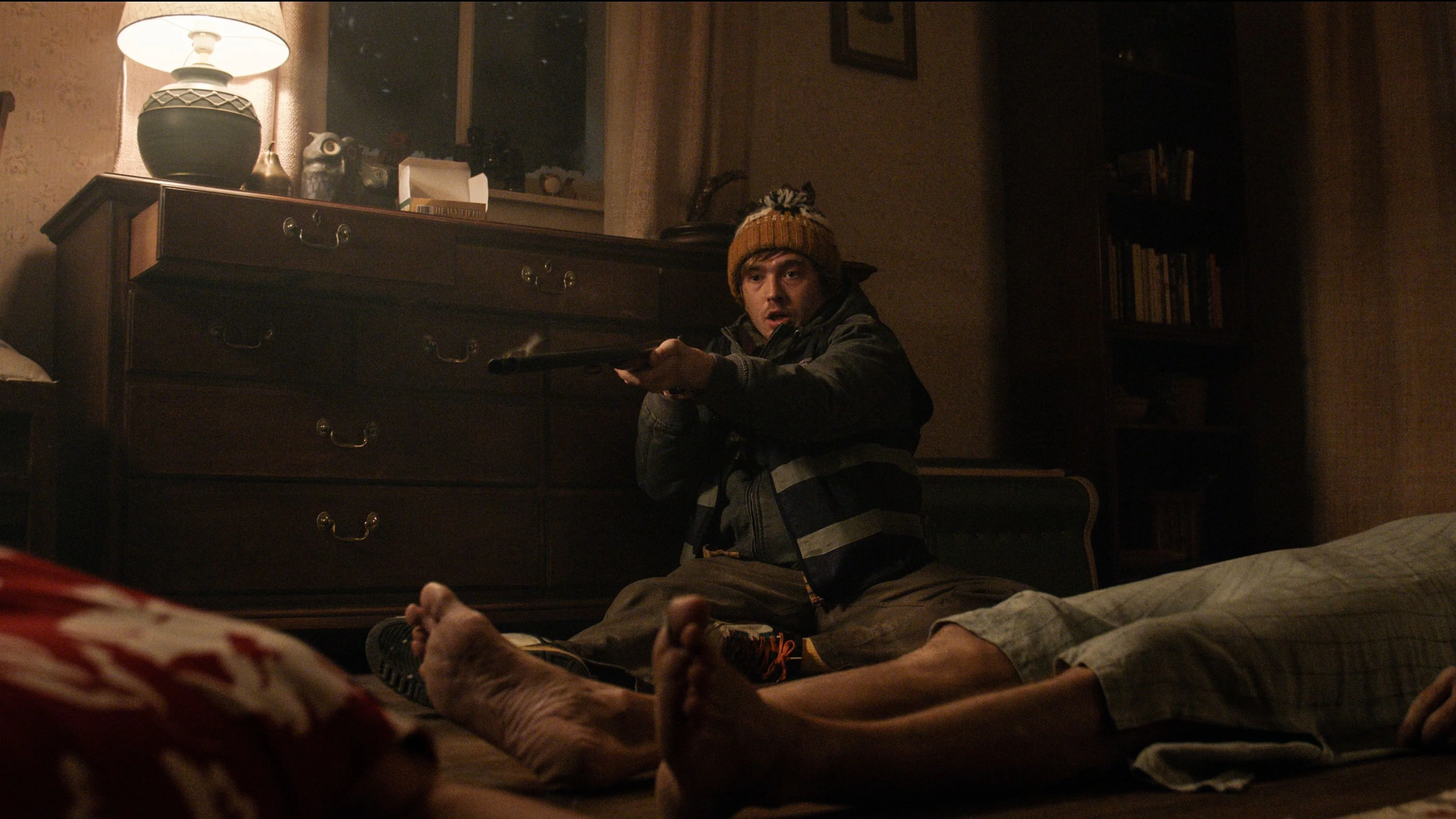 Bryan sits in a bedroom and aims a shotgun with both hands.