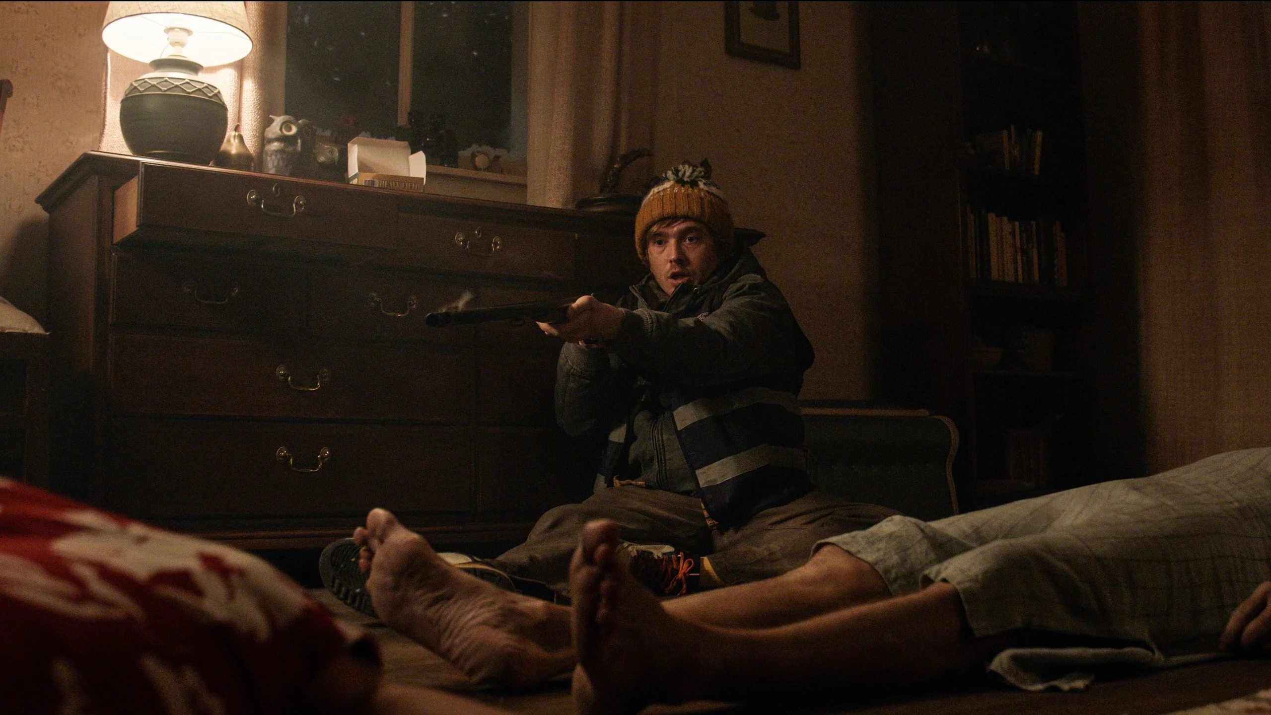 Bryan sits in a bedroom and aims a shotgun with both hands.