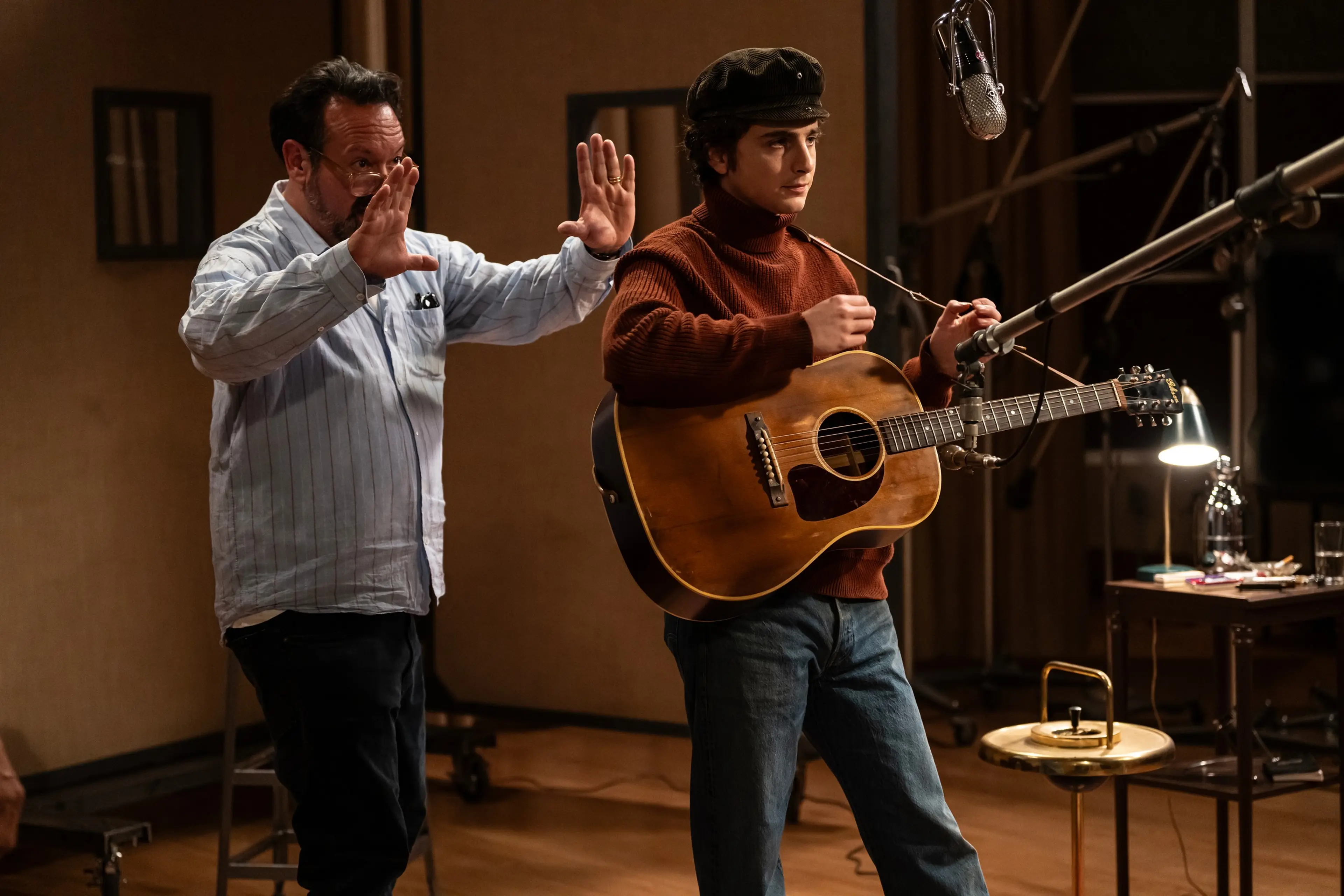 Director James Mangold gestures beside Timothée Chalamet, who is holding an acoustic guitar near microphones.