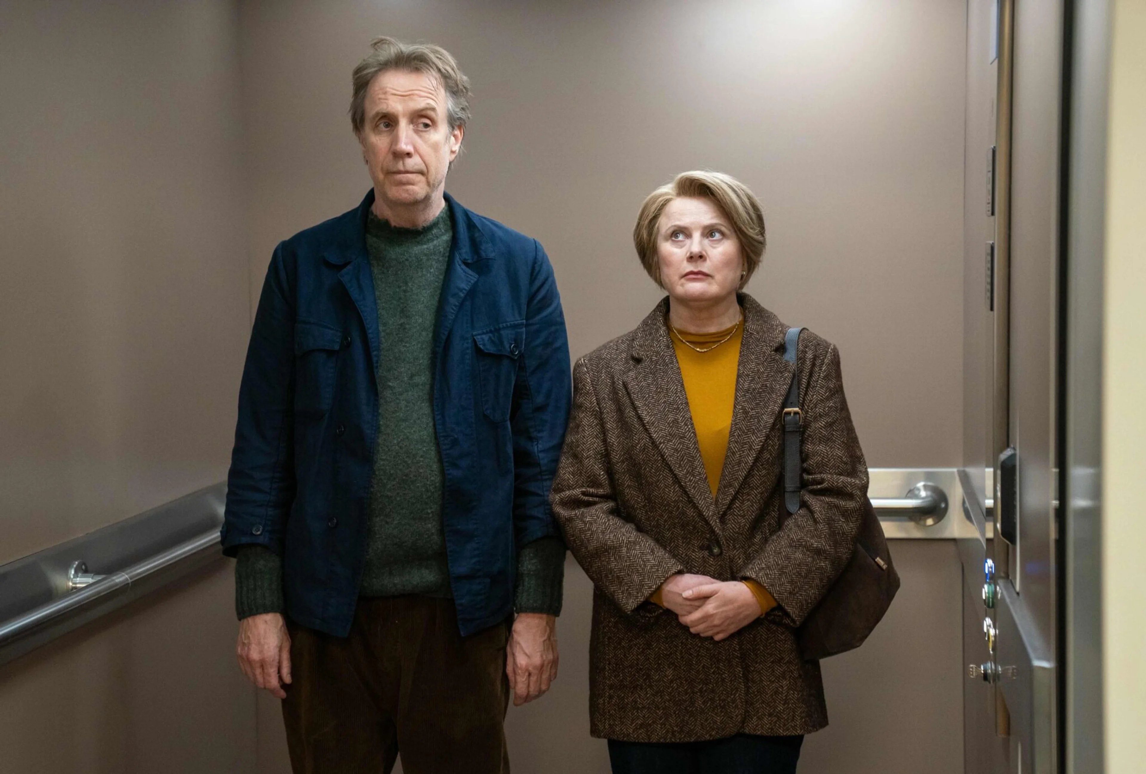 Rhys Ifans as Tony and Laura Linney as Emma stand together in an elevator.
