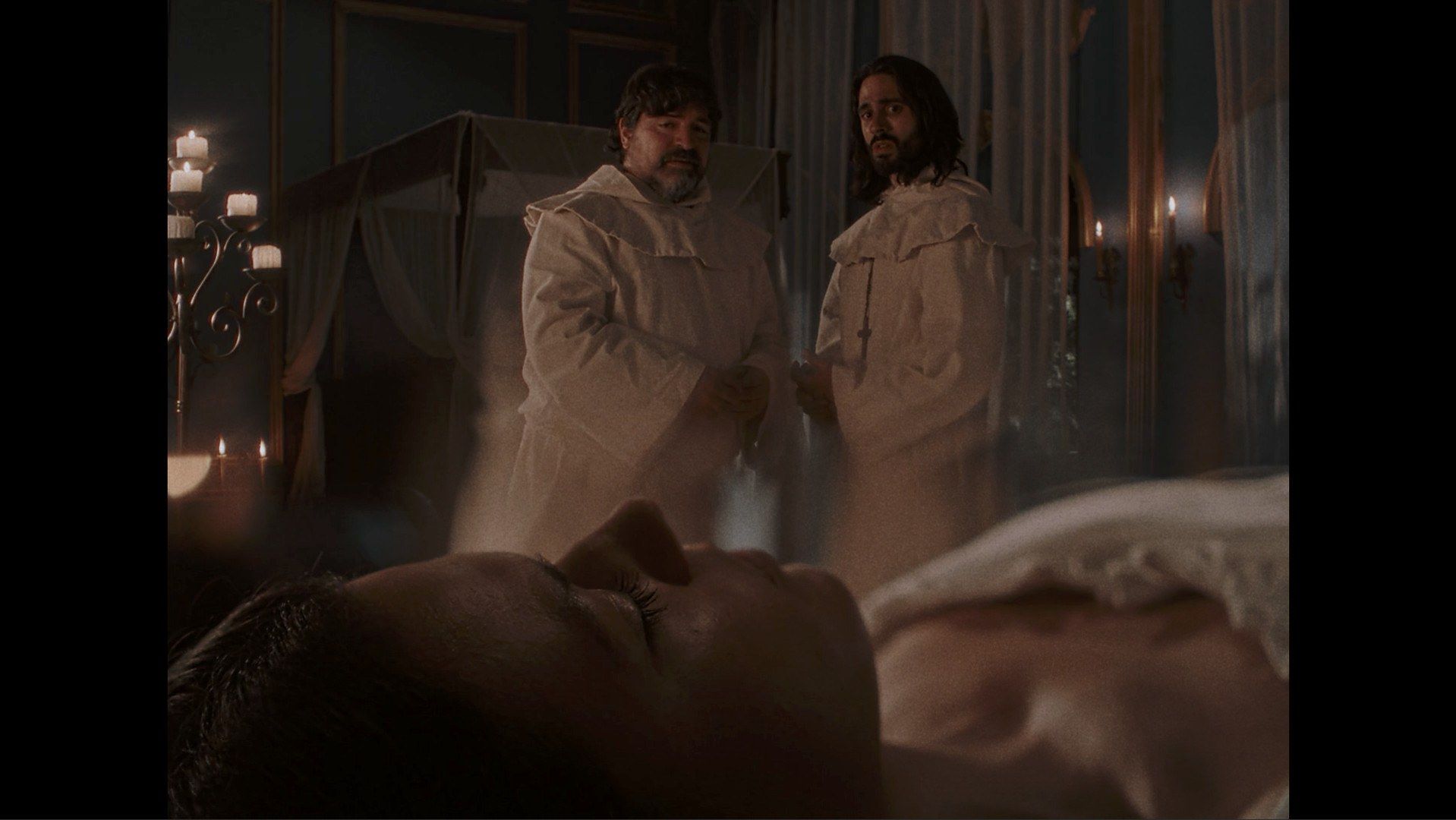 Two actors stand above a person lying on a bed in a film scene.
