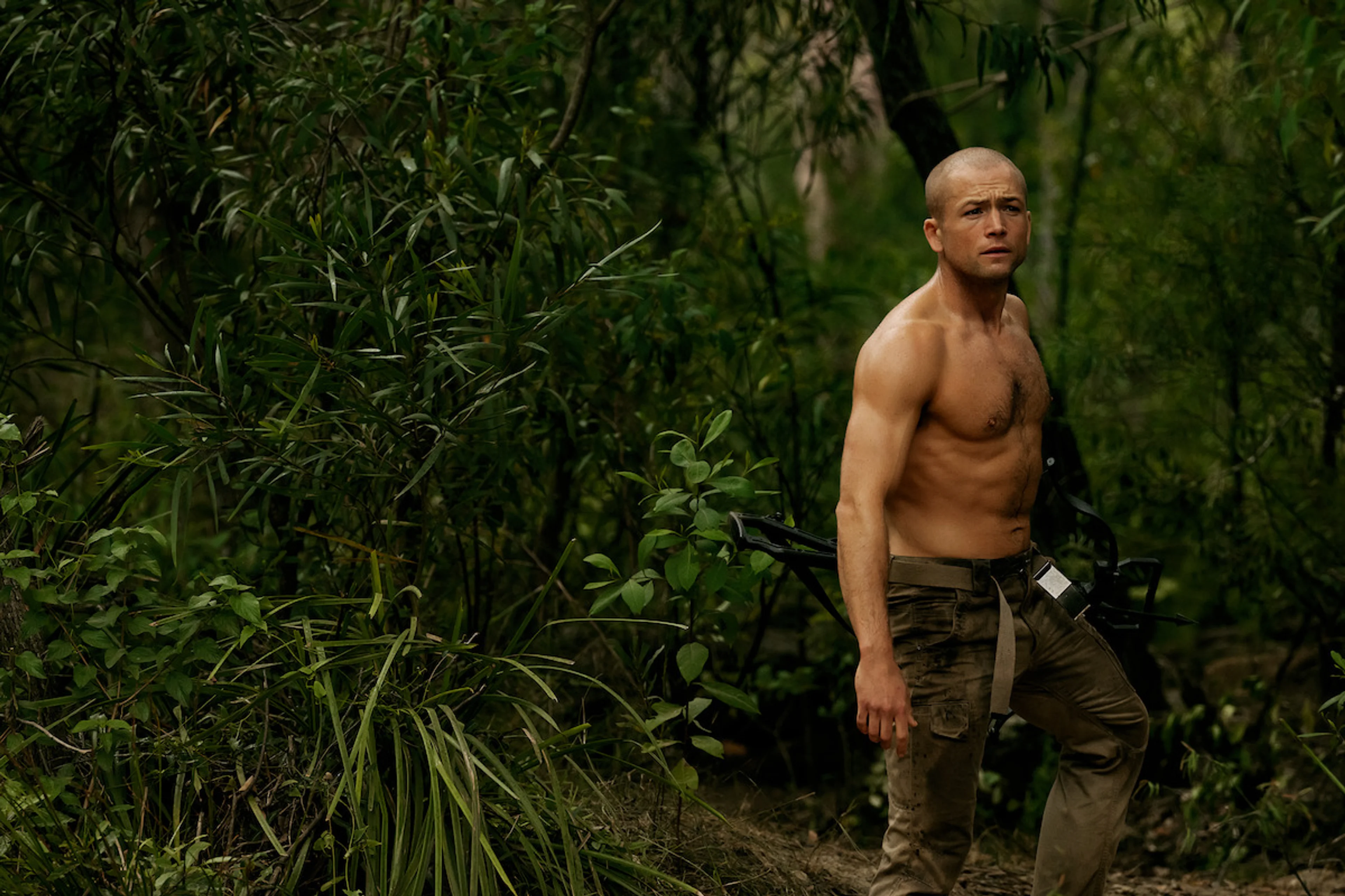 Taron Egerton as Ben stands in the forest carrying a crossbow.