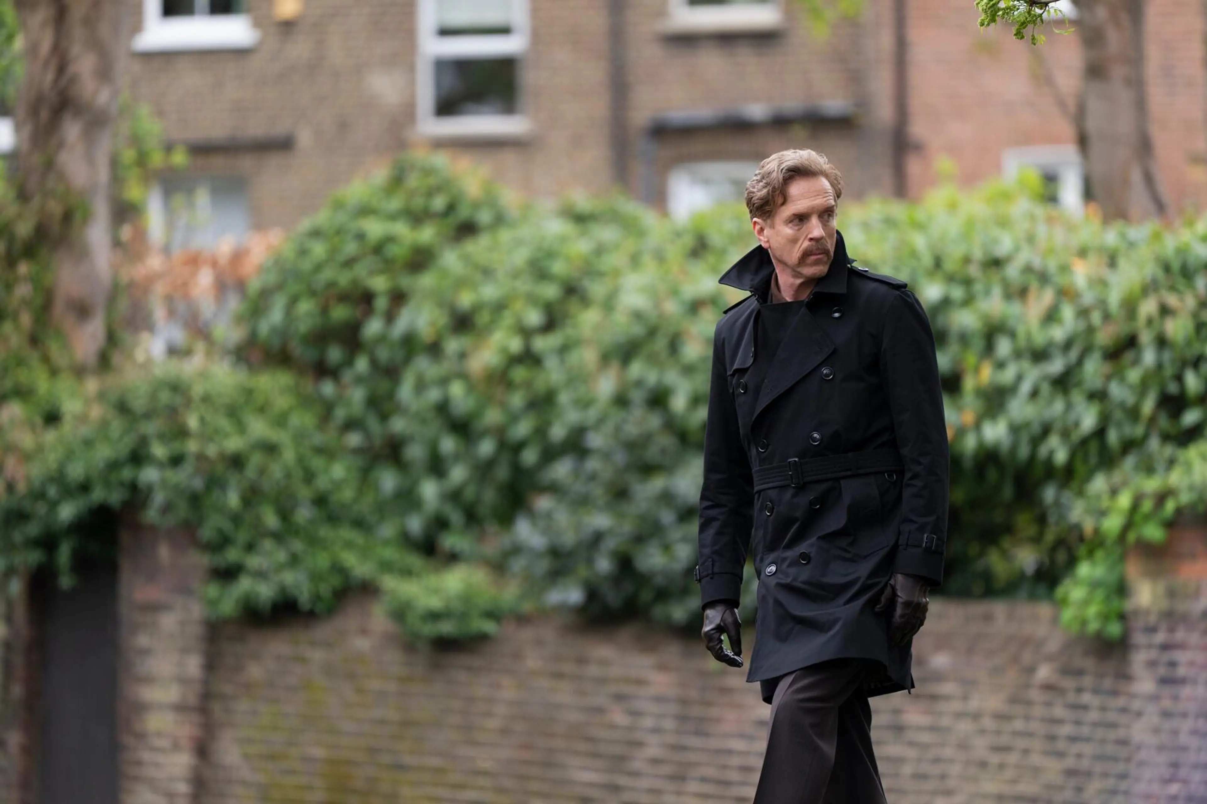 Damian Lewis as the Caller walks beside a brick building in The Runner.