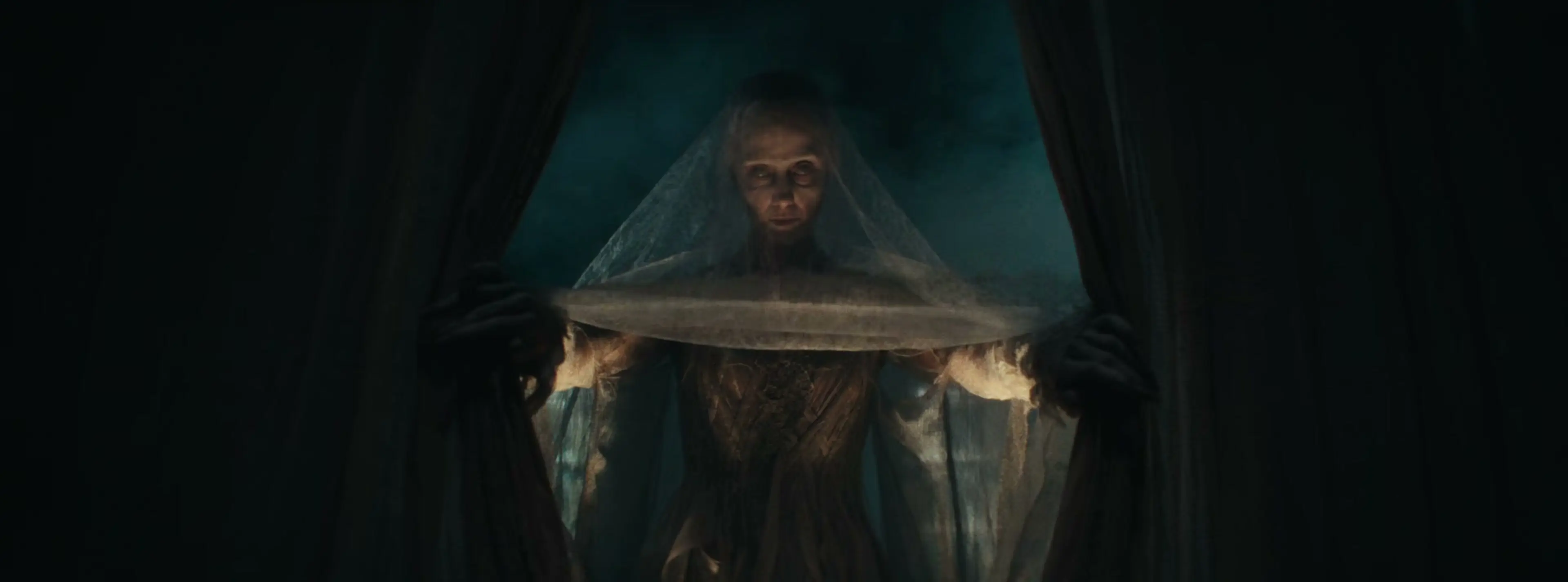 A pale veiled figure stands between the curtains of Scrooge’s bed.