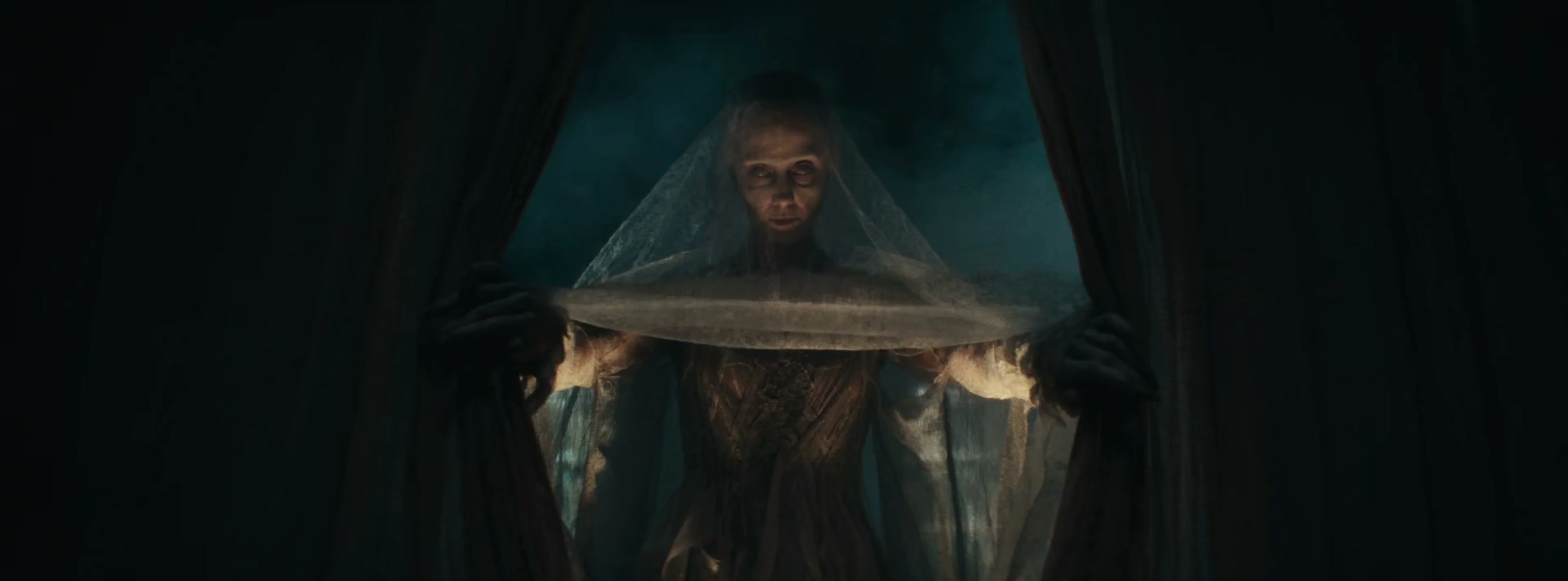 A pale veiled figure stands between the curtains of Scrooge’s bed.