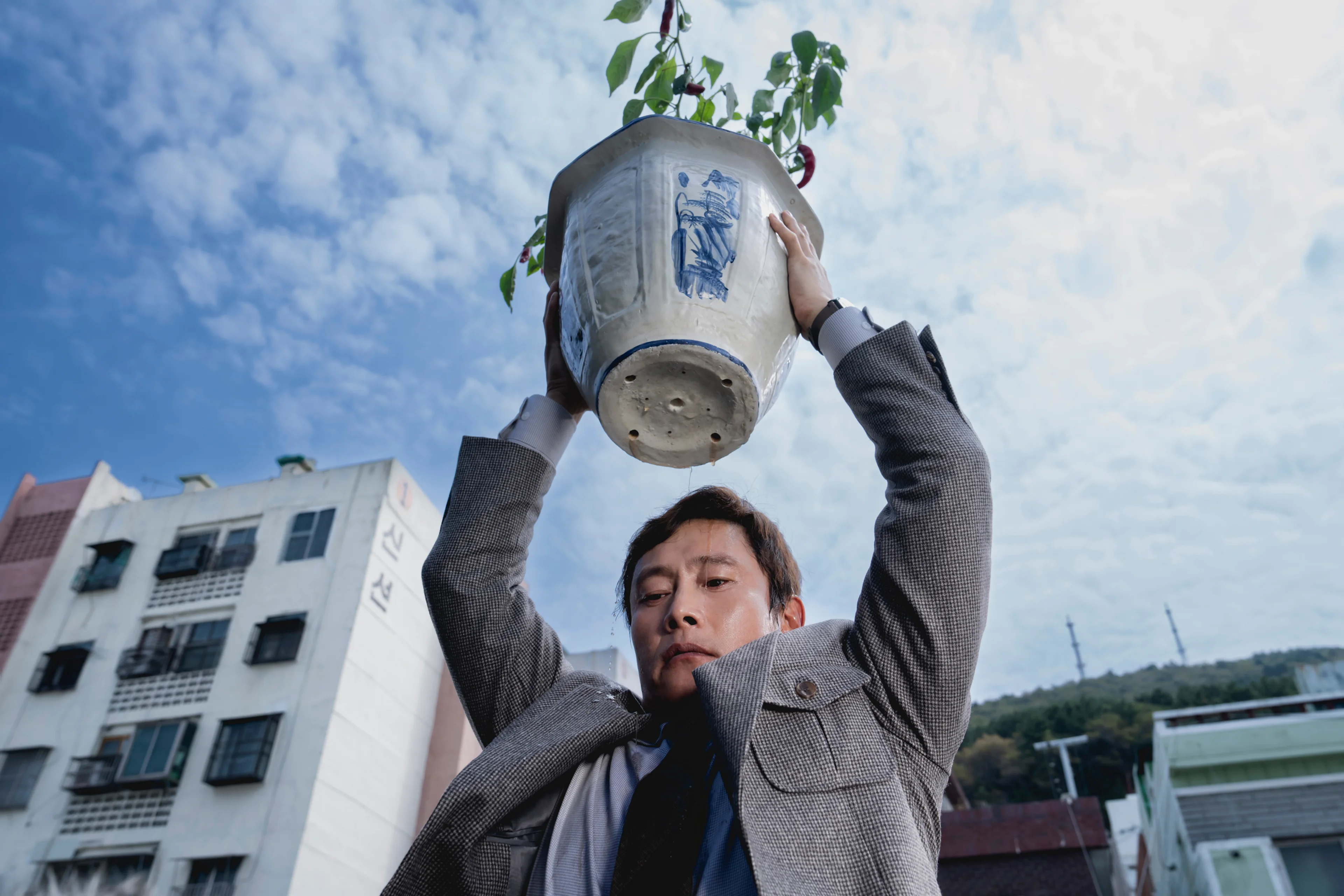 Man-su lifts a large plant pot above his head outdoors.