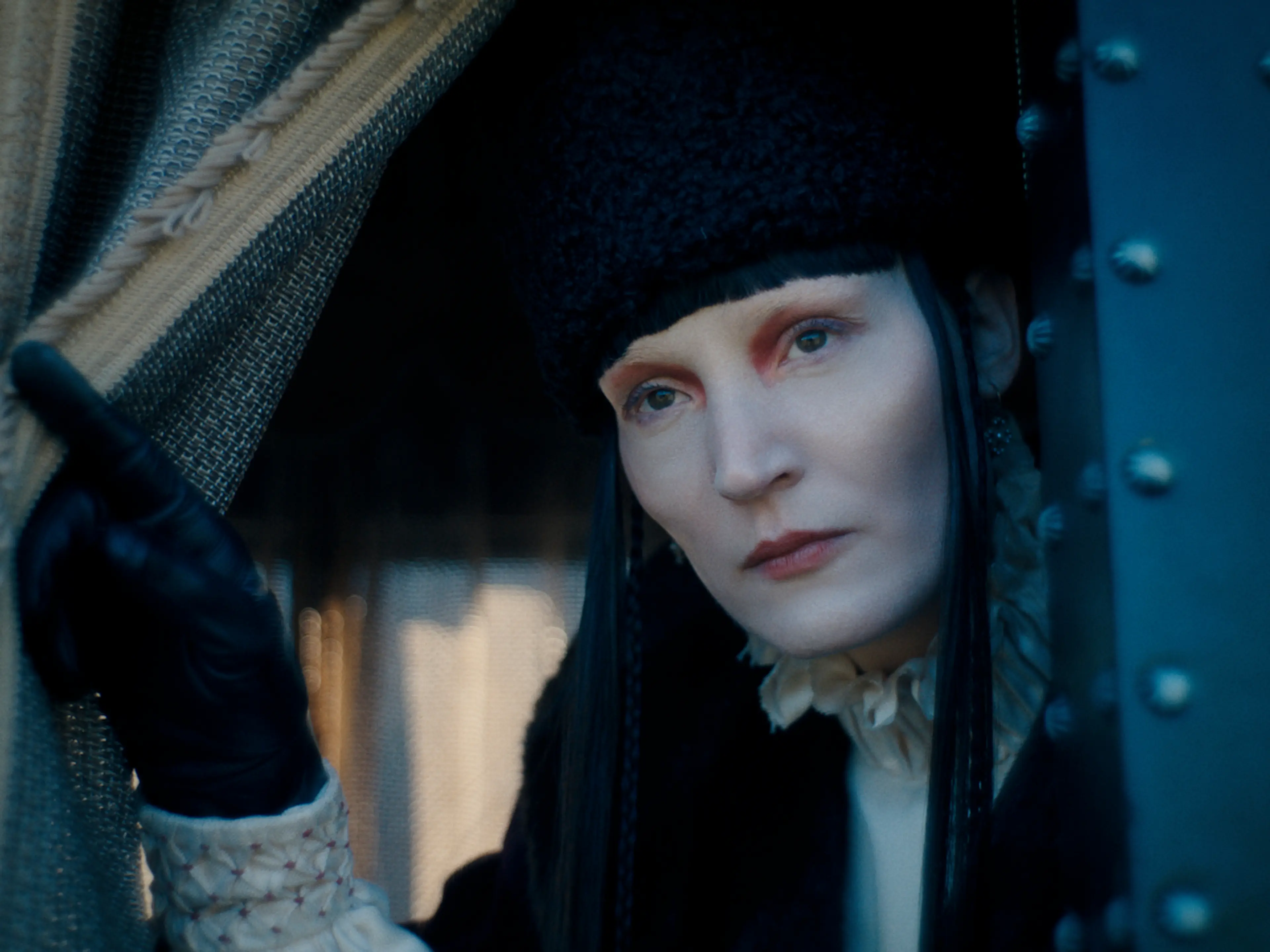 Vicky Krieps as Elizabeth Báthory wears a black fur hat and gloves beside a curtain.