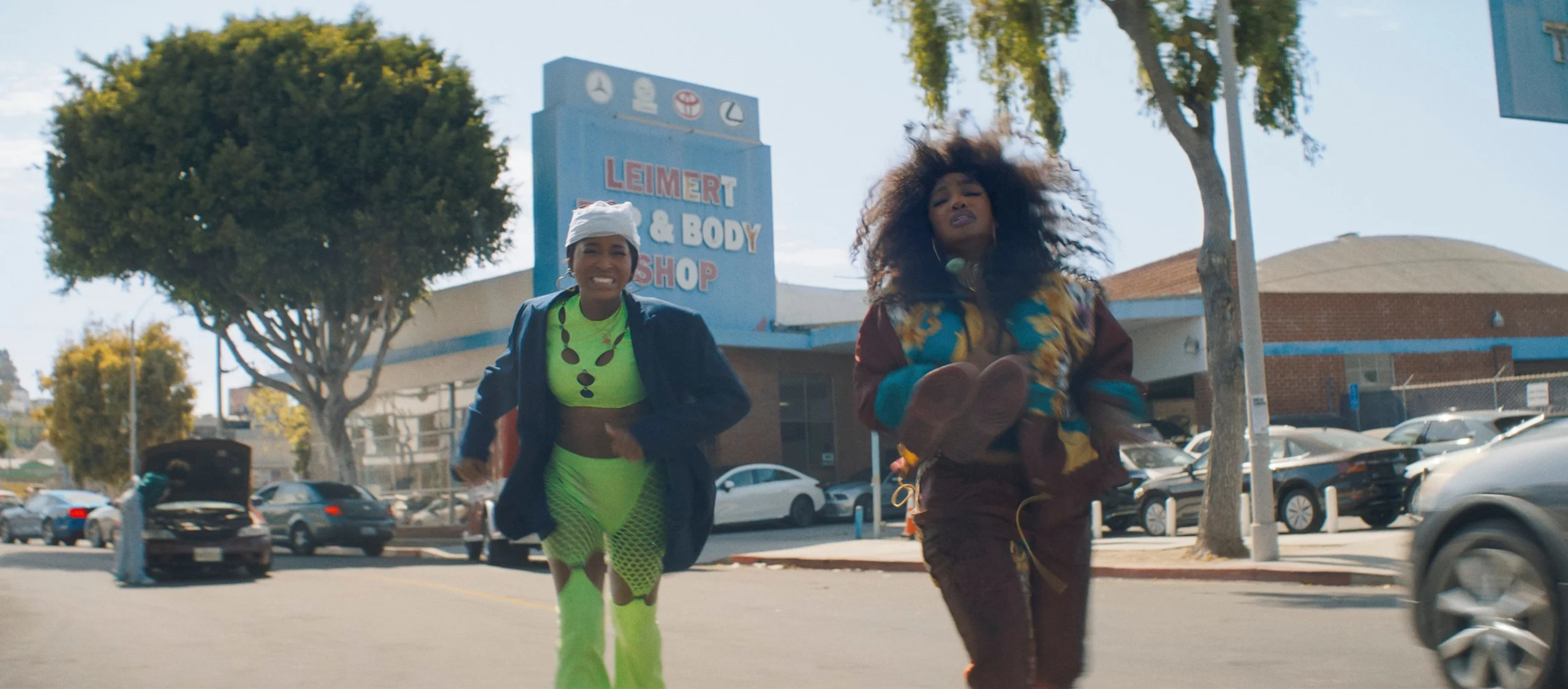 Keke Palmer in bright green clothes and SZA in a patterned top run along a sunny Los Angeles street.