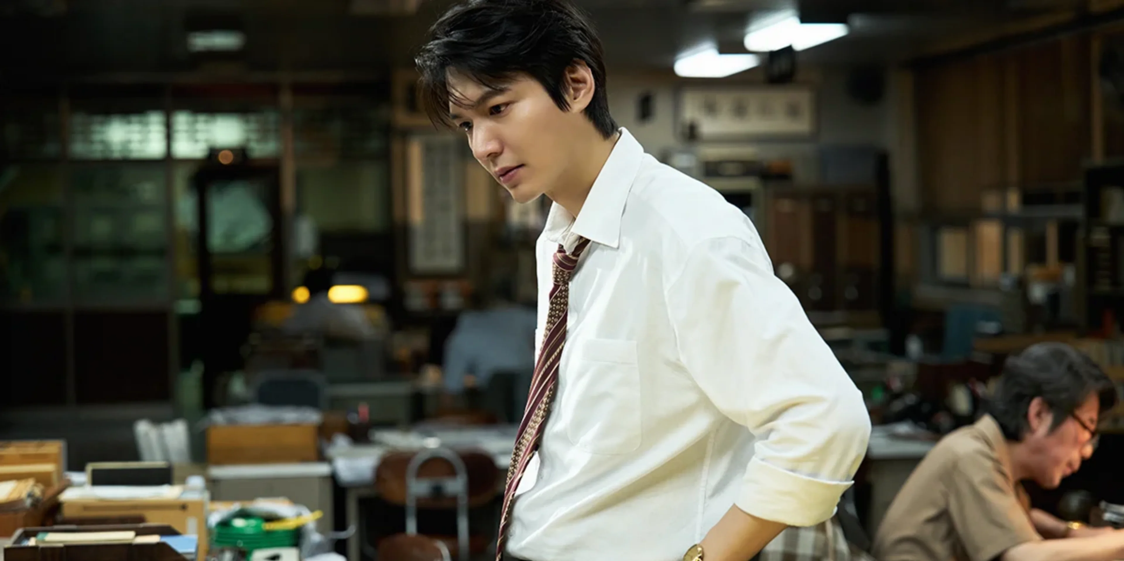 Lee Min-ho as reporter Young-il leans over a desk in the newspaper office.
