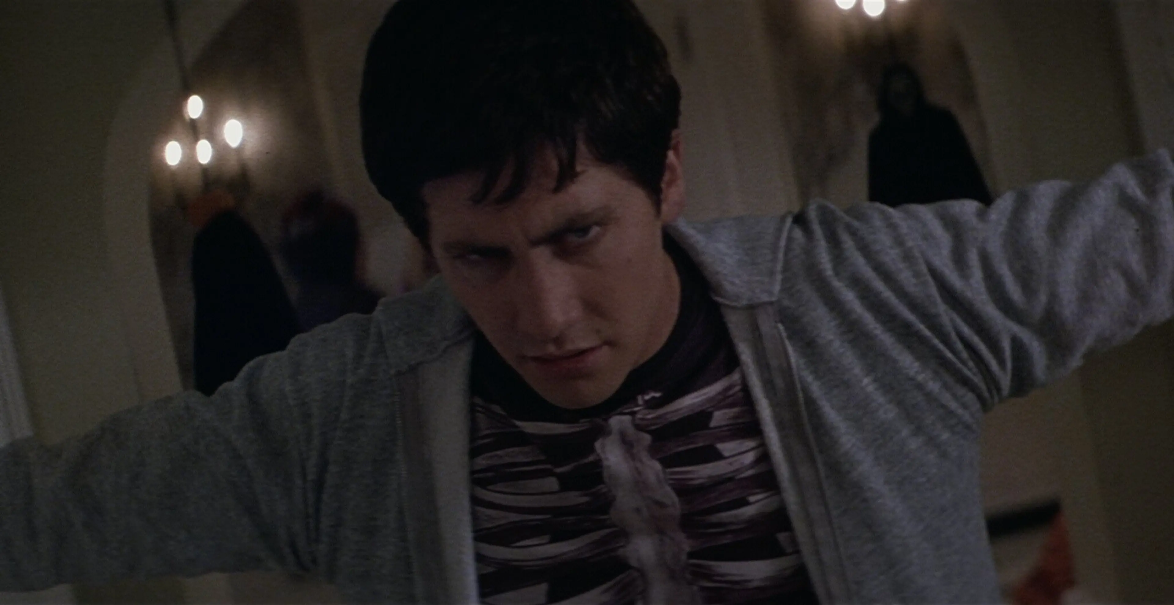 Jake Gyllenhaal as Donnie Darko leans forward in a gray jacket and striped shirt.