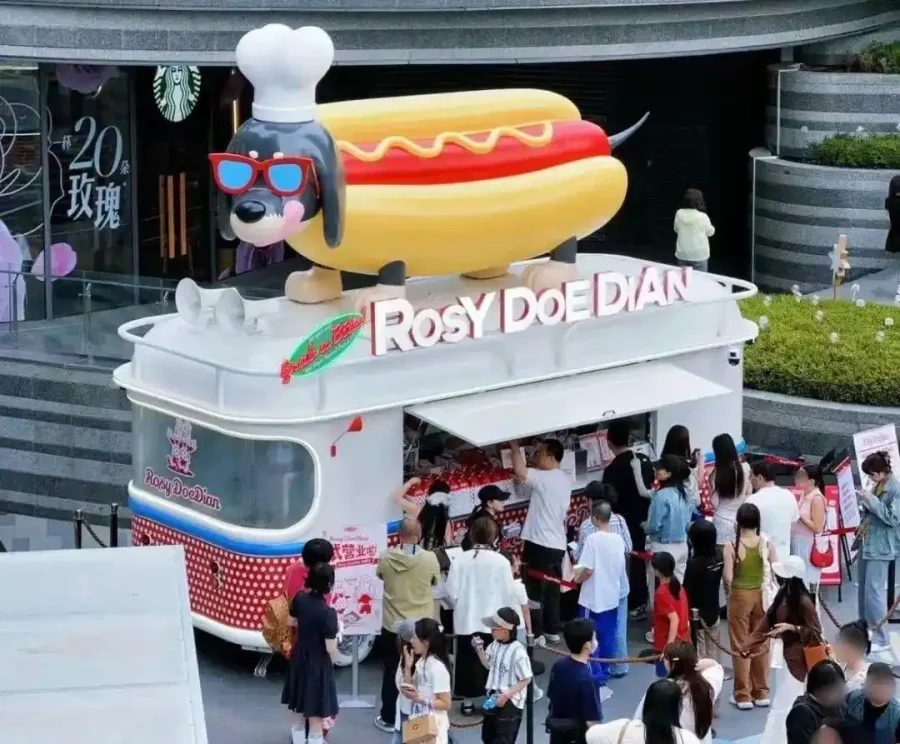 Rosy DoeDian mobile hot dog cart beneath a dachshund sculpture, with customers waiting nearby.