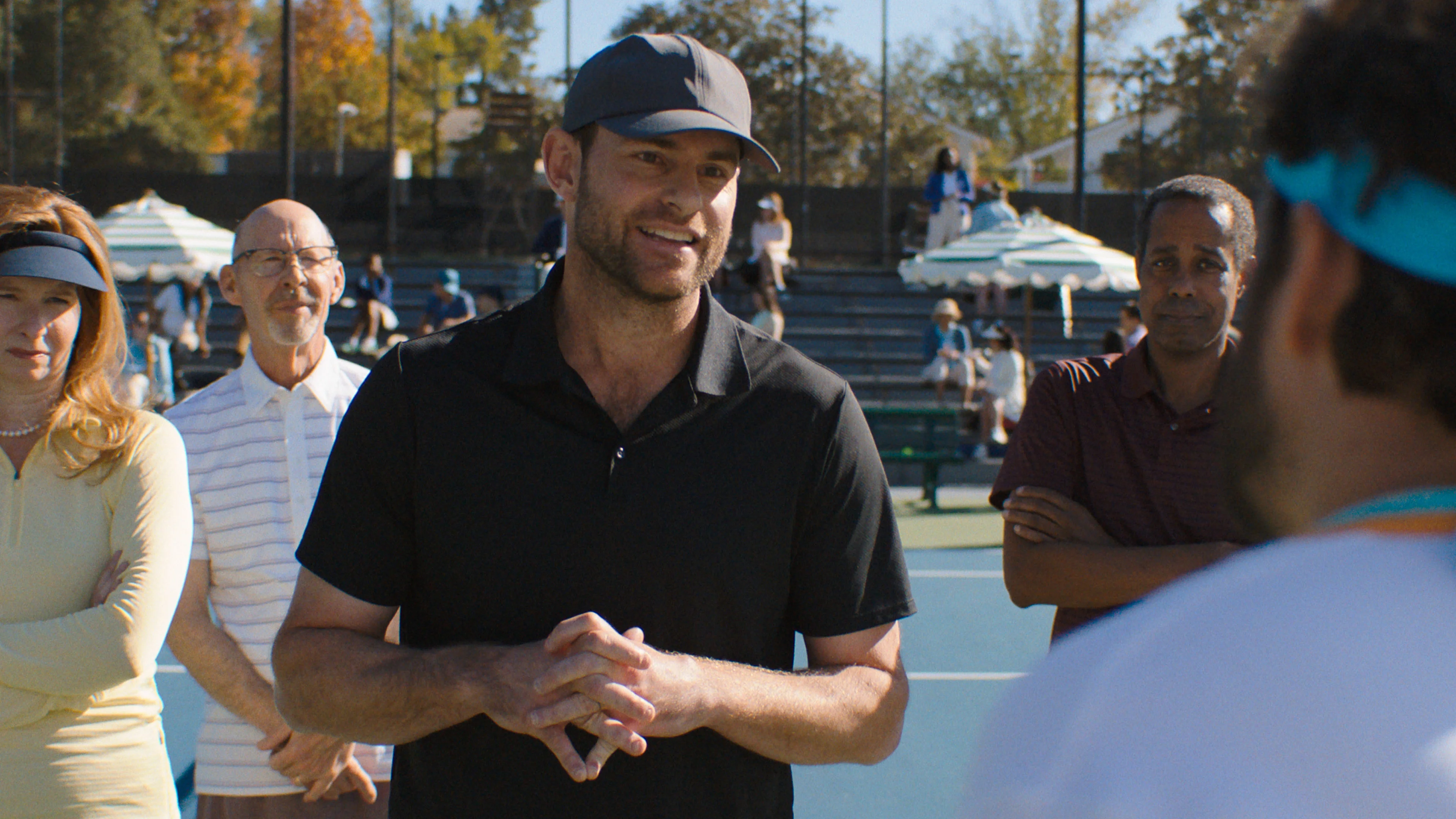 Andy Roddick wears a black shirt and cap while speaking to Dusty on a court.
