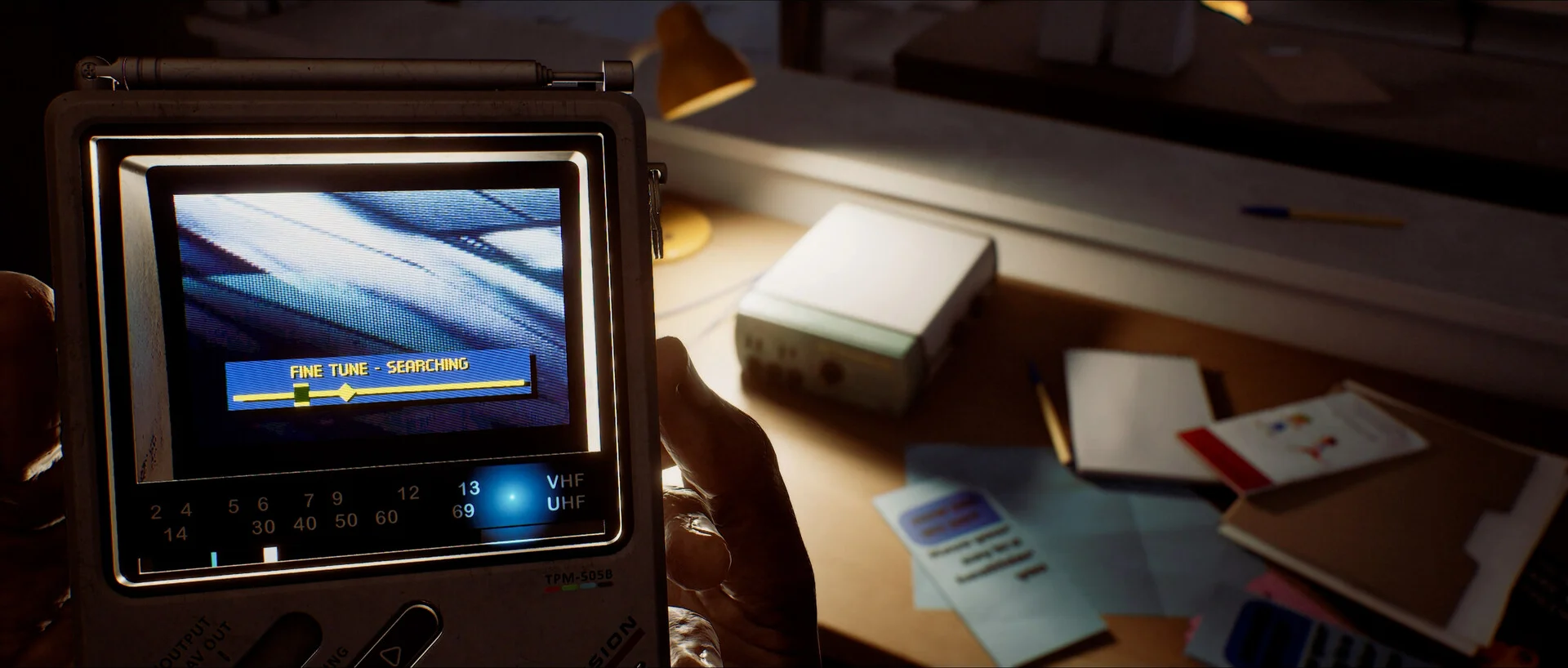 A hand holds a pocket television tuned towards desks and papers inside a building.