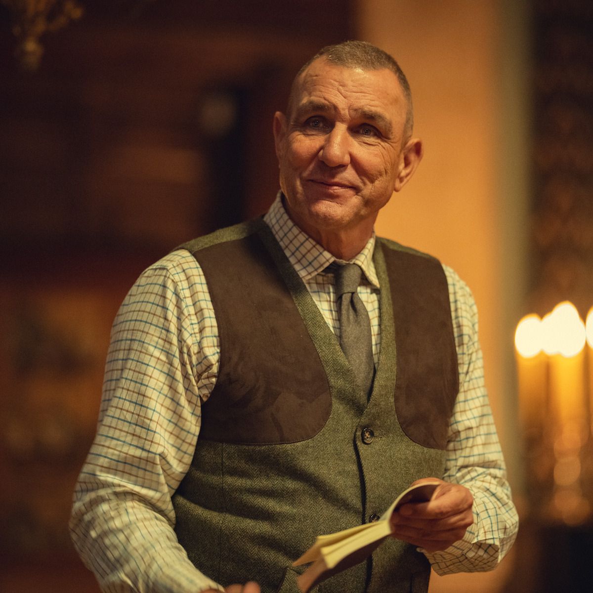 Vinnie Jones as Geoff Seacombe holds an open book while wearing a checked shirt and waistcoat.