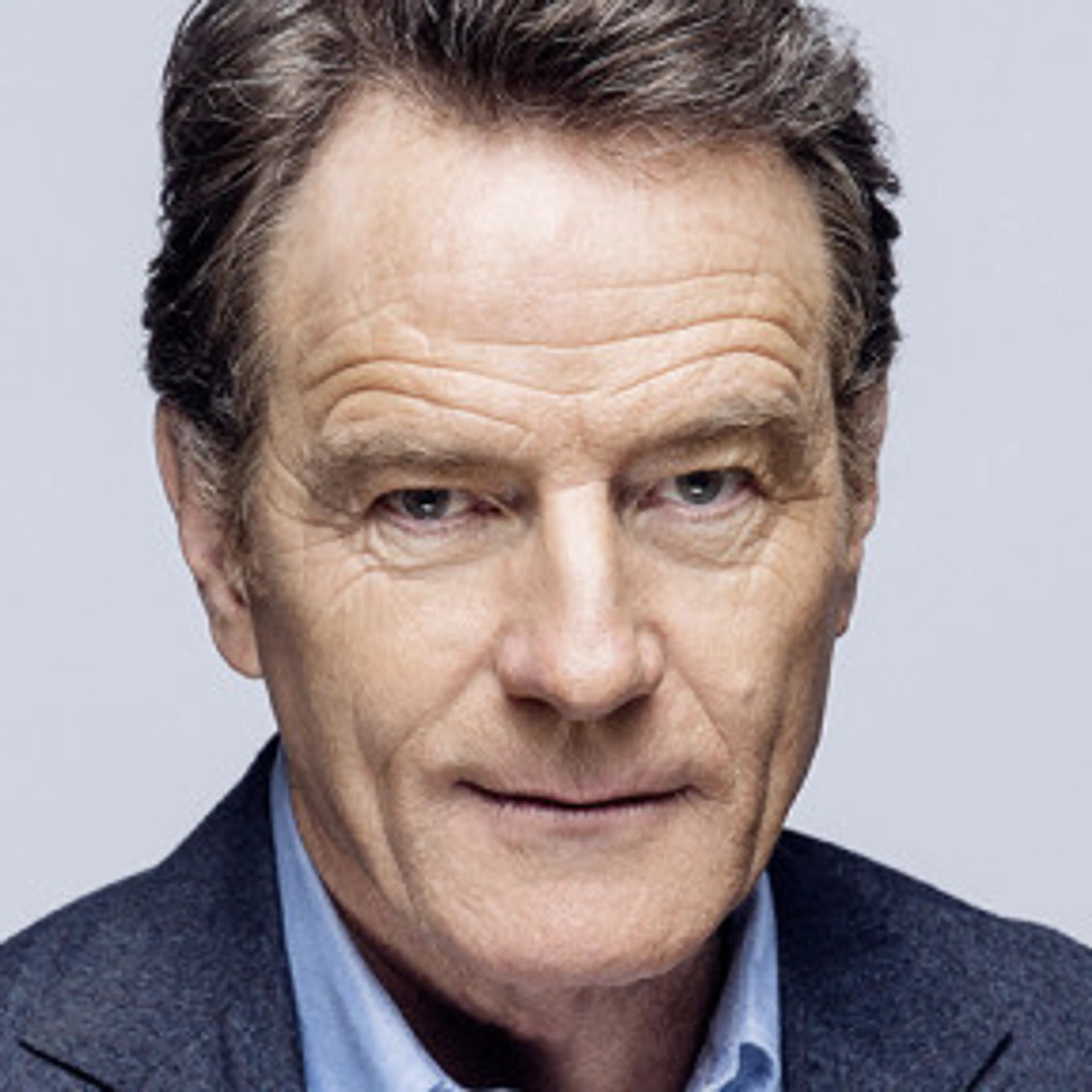Bryan Cranston in a dark blazer and blue shirt against a pale gray background.