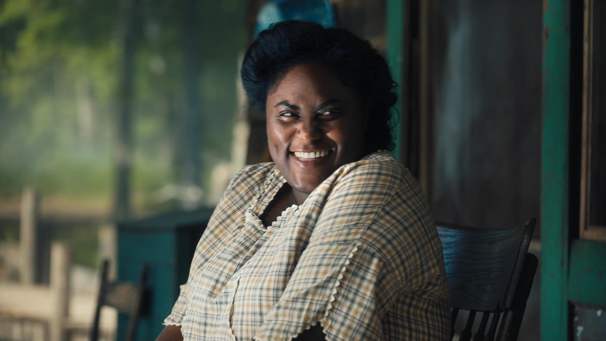 Danielle Brooks as Sofia smiles while seated in a wooden chair in The Color Purple (2023).