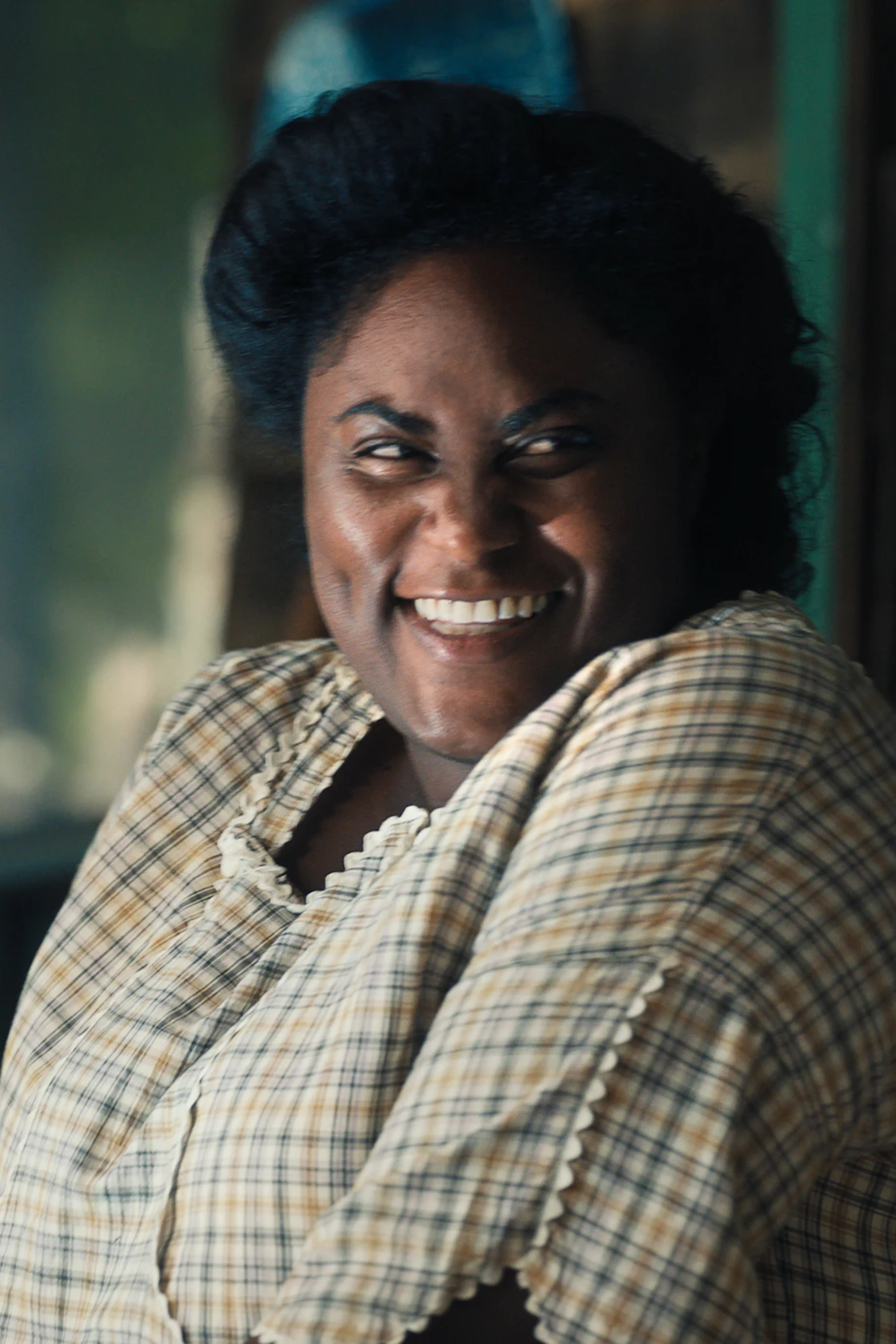 Danielle Brooks as Sofia smiles while seated in a wooden chair in The Color Purple (2023).