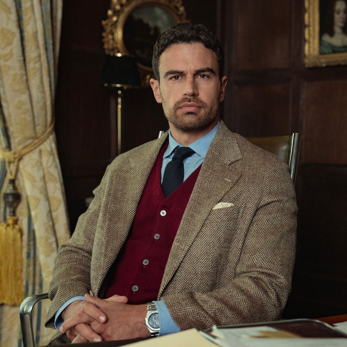 Theo James as Eddie Horniman seated in a wood-paneled office wearing a brown suit.