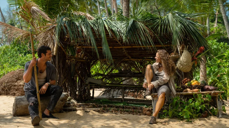 Bradley and Linda sit at either side of a palm-leaf shelter with food stacked nearby.