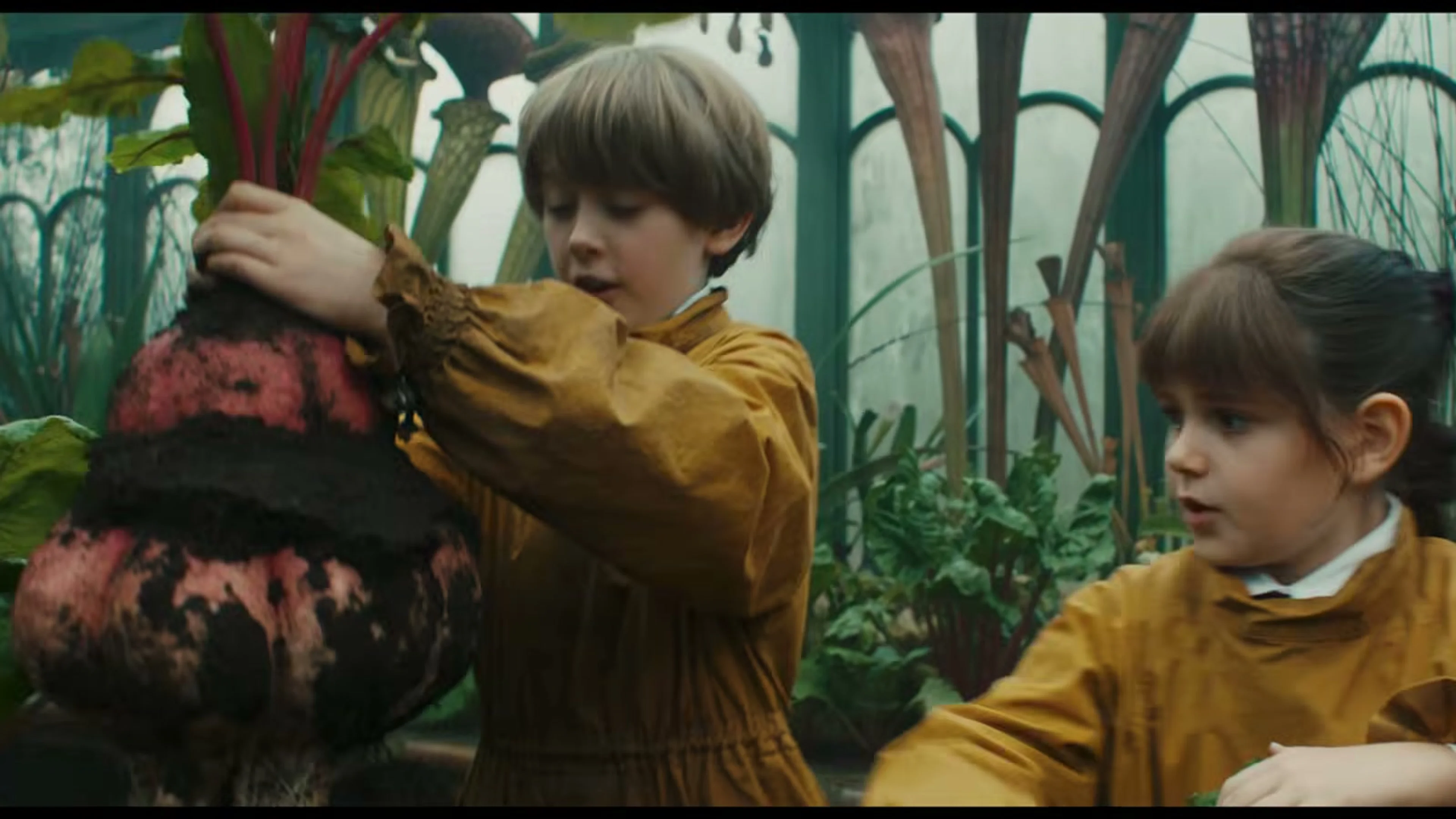 Two Hogwarts students handle a large red-and-black magical plant in a greenhouse.