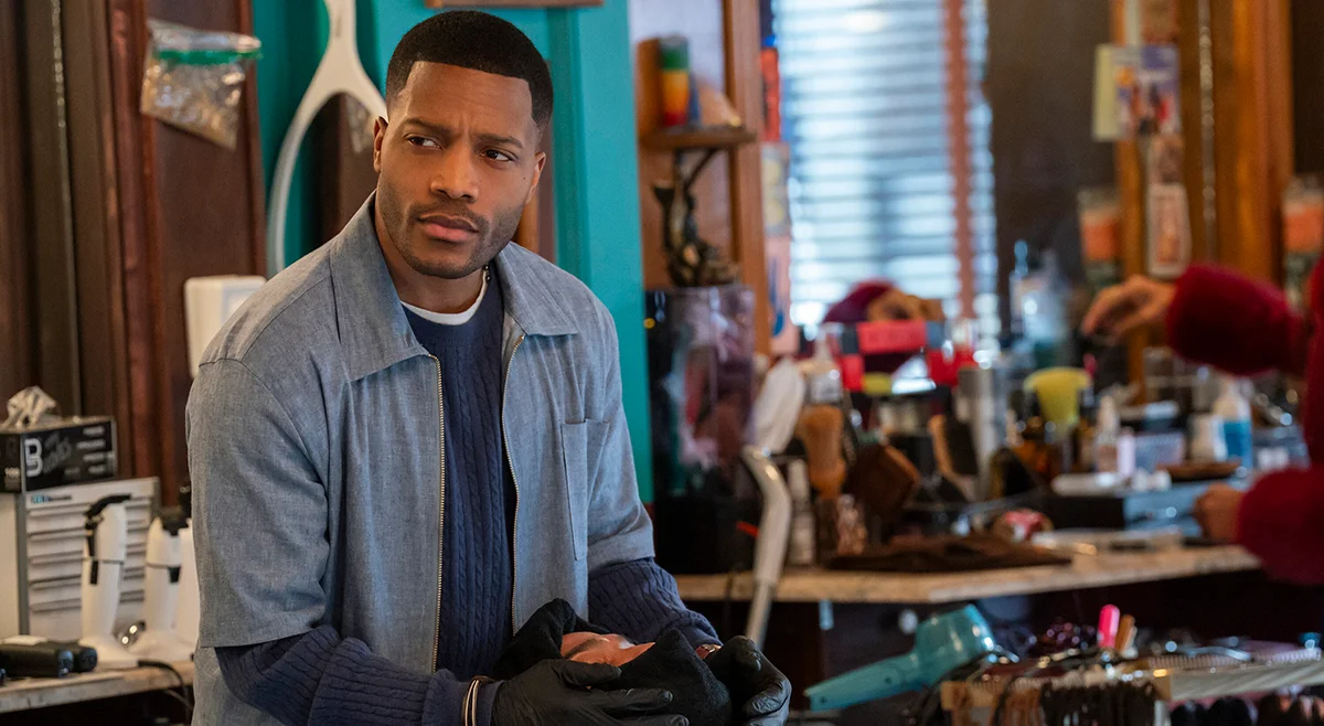 Jermaine Fowler wears a blue shirt and holds a dark jacket inside the barbershop.