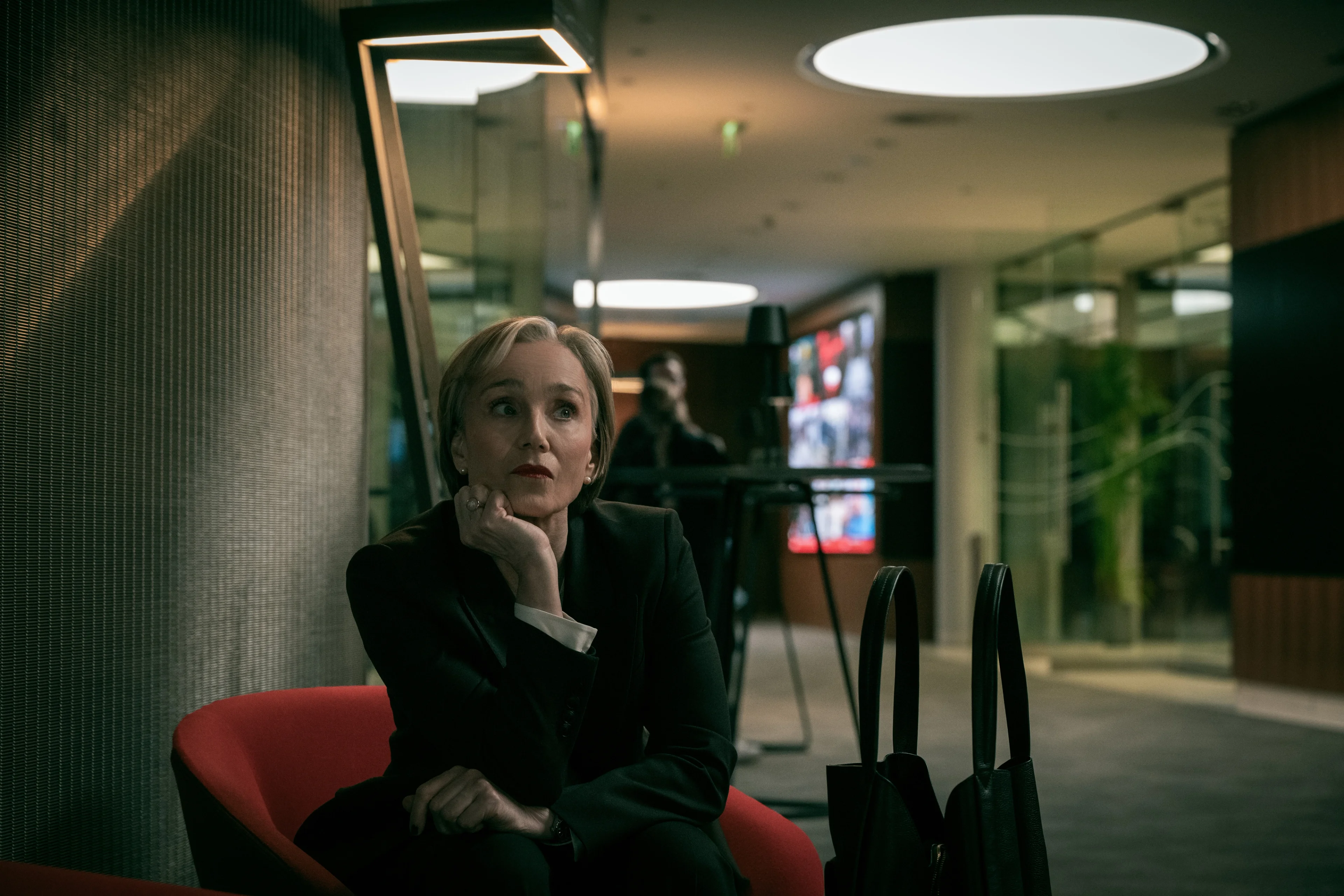 Diana Taverner sits in a red chair with her chin resting on her hand.