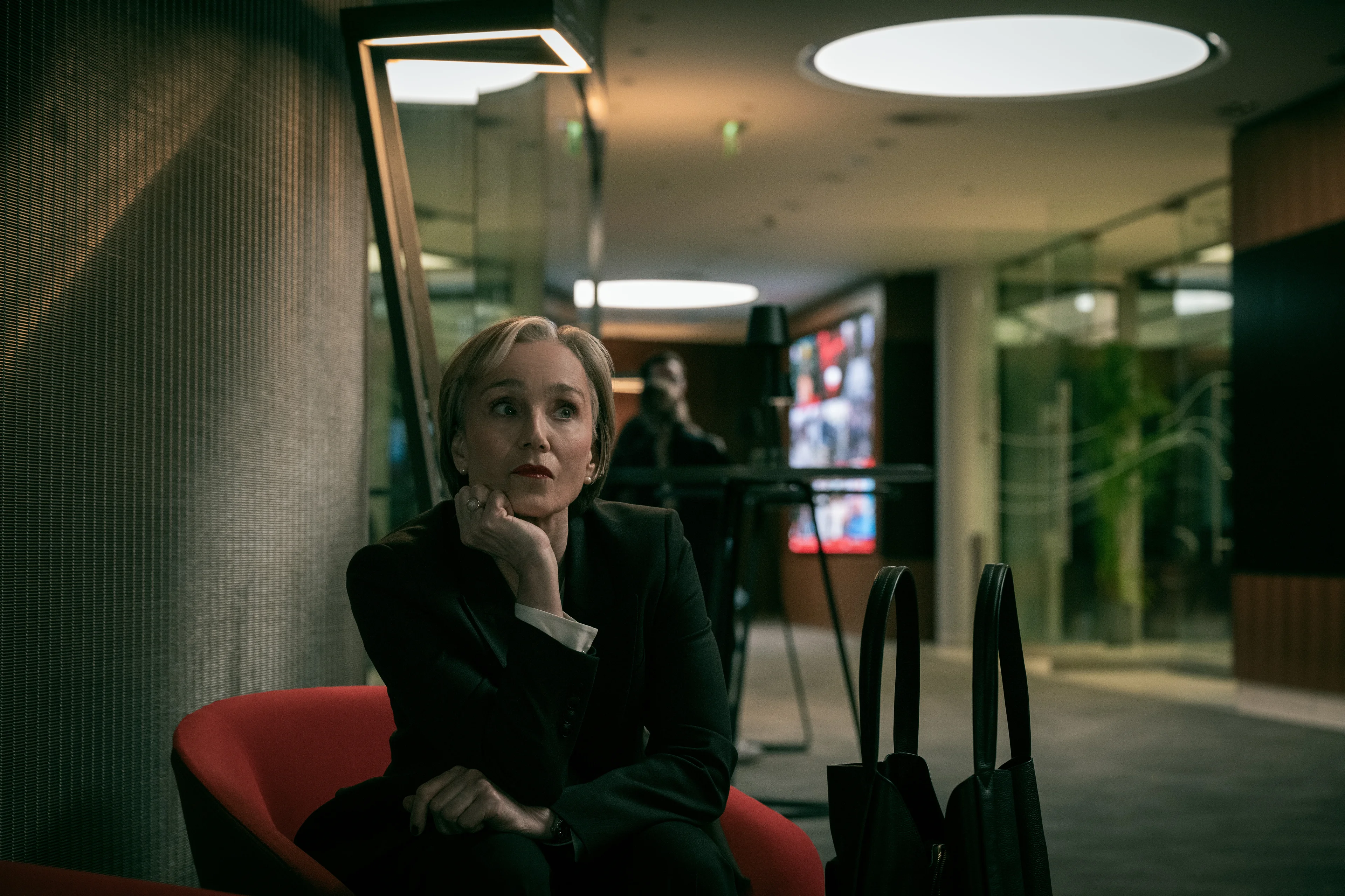 Diana Taverner sits in a red chair with her chin resting on her hand.