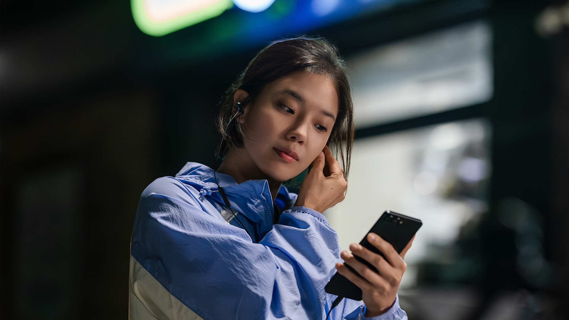 A runner in a blue jacket adjusts her earphone while holding a phone in Your Personal Taxi.