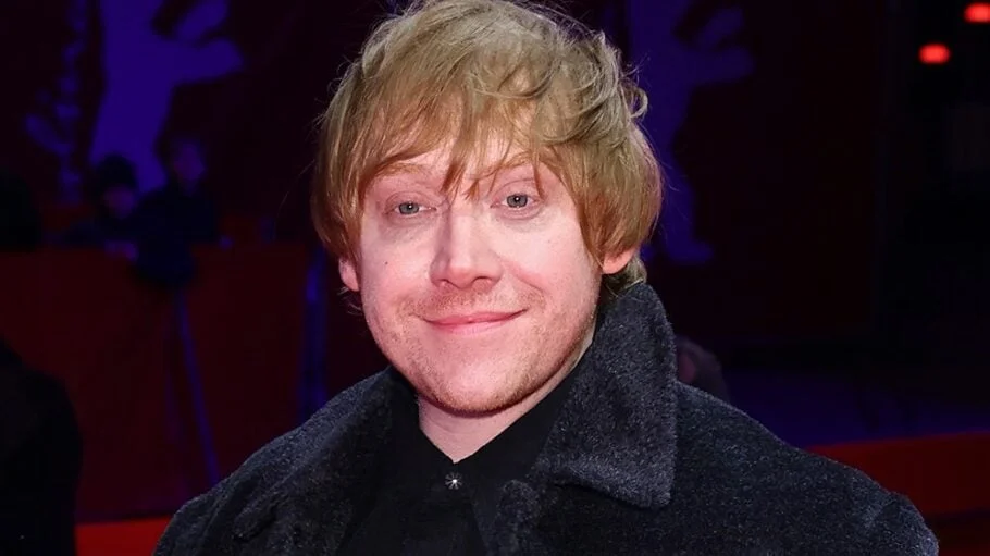 Rupert Grint smiles at a premiere in a dark coat.