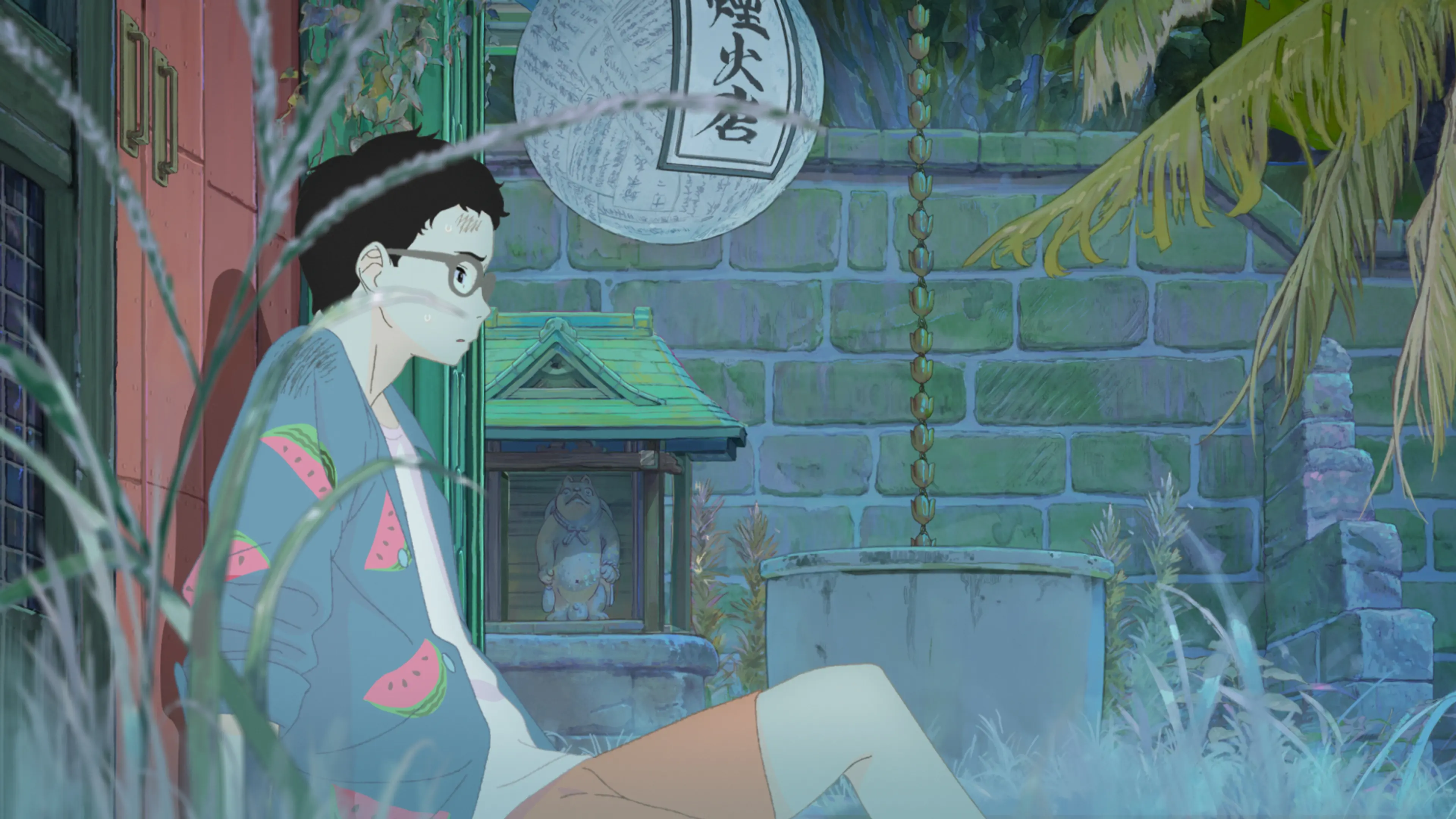A young man with glasses sits outdoors in a watermelon-patterned robe beside a bucket and greenery.