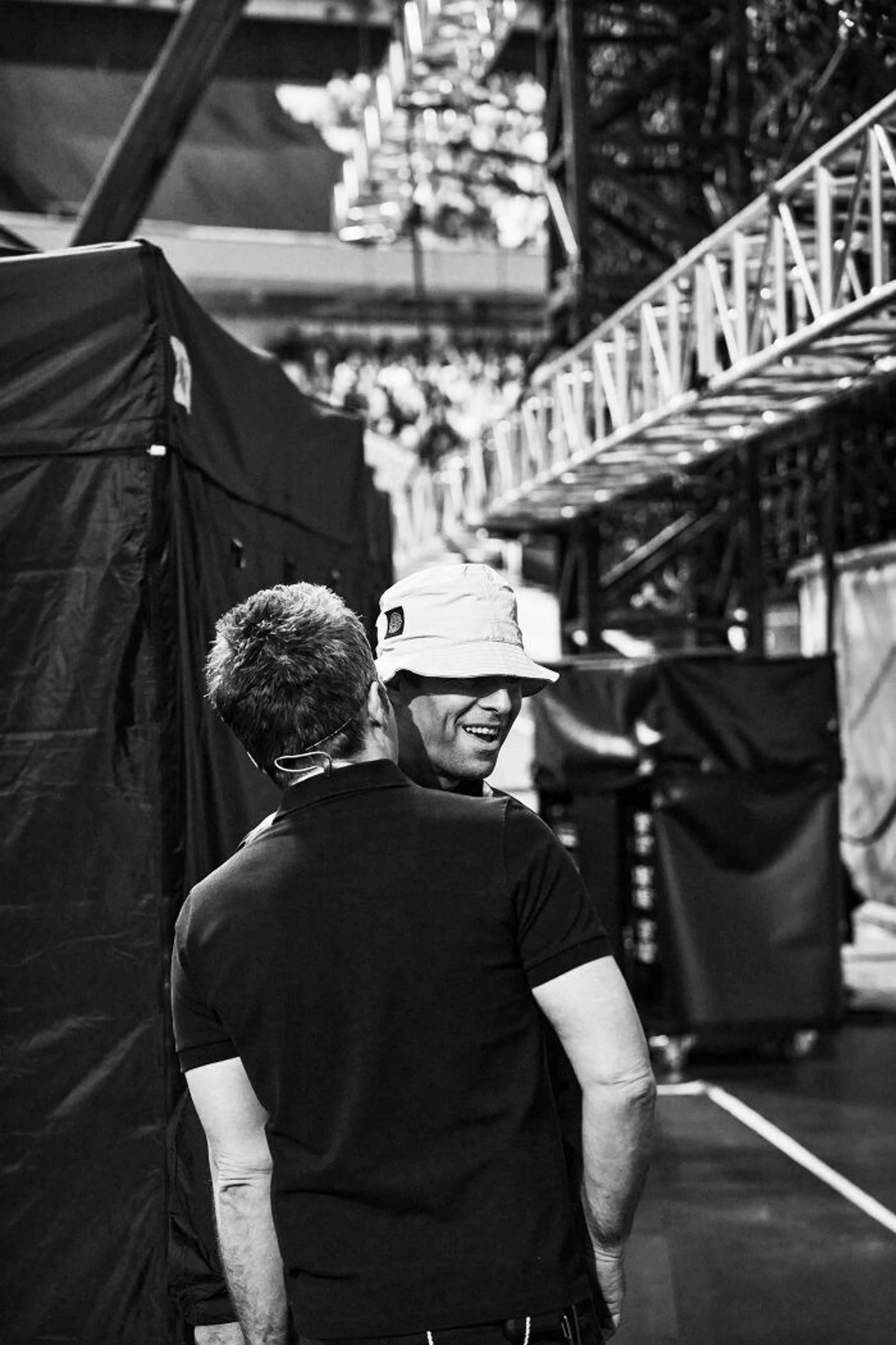 The Gallagher brothers embrace backstage in a black-and-white vertical documentary still.