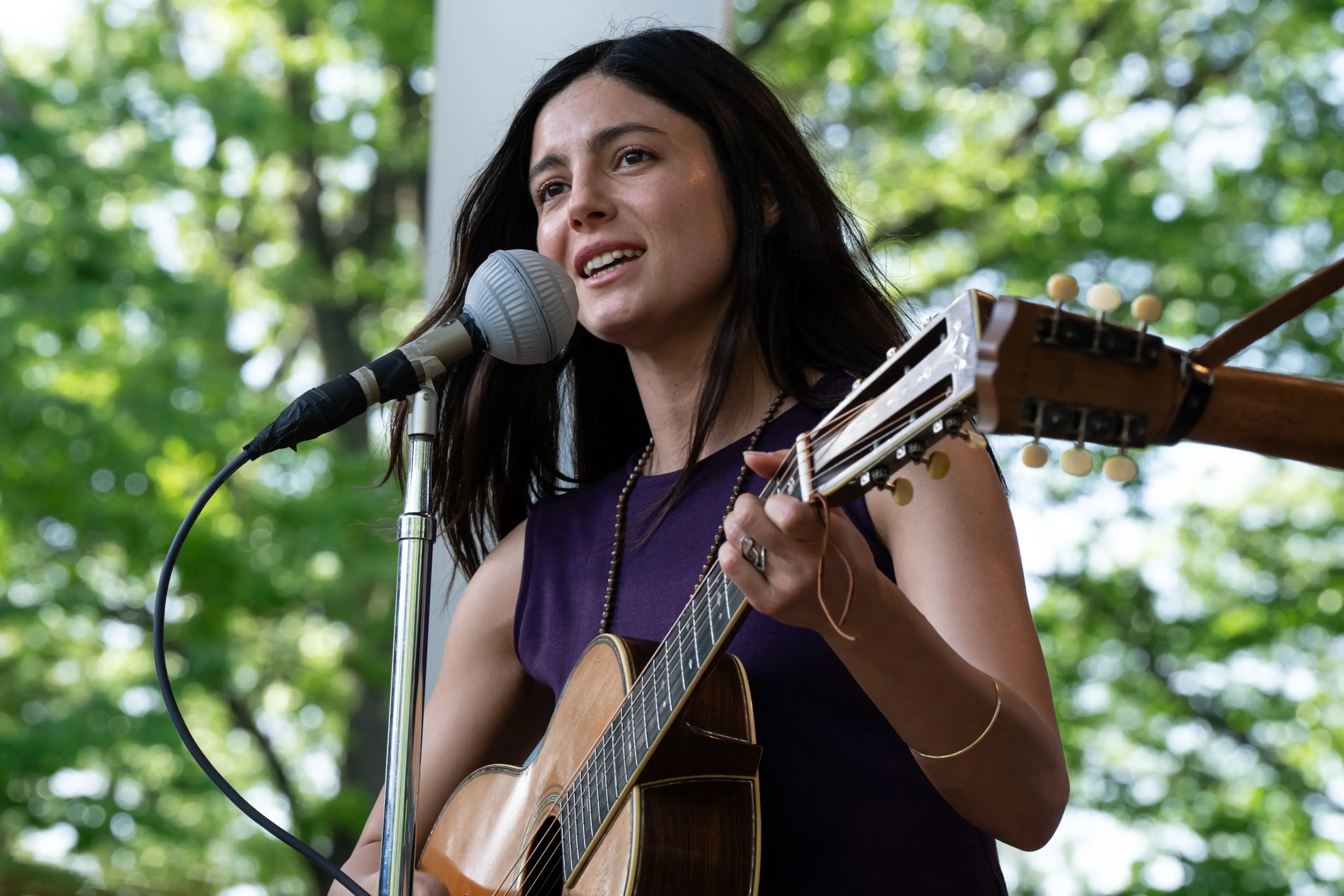 Monica Barbaro as Joan Baez sings into a microphone while playing guitar outdoors.