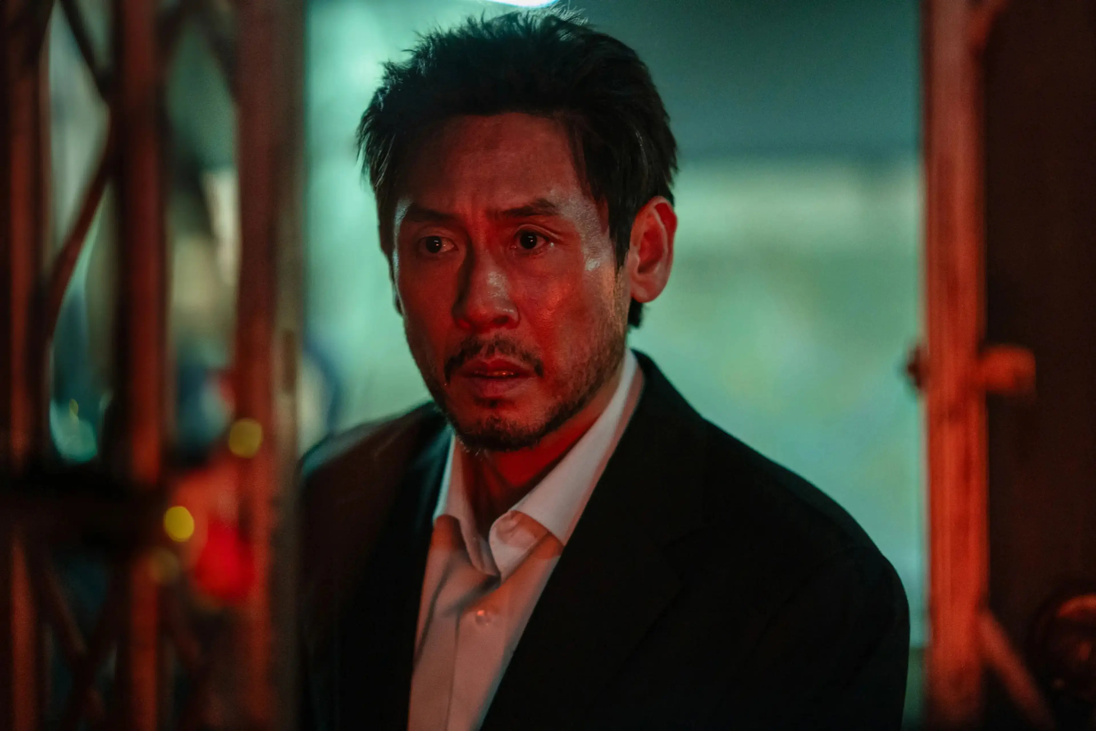 Noja looks ahead while wearing a dark suit and an open-collared white shirt.
