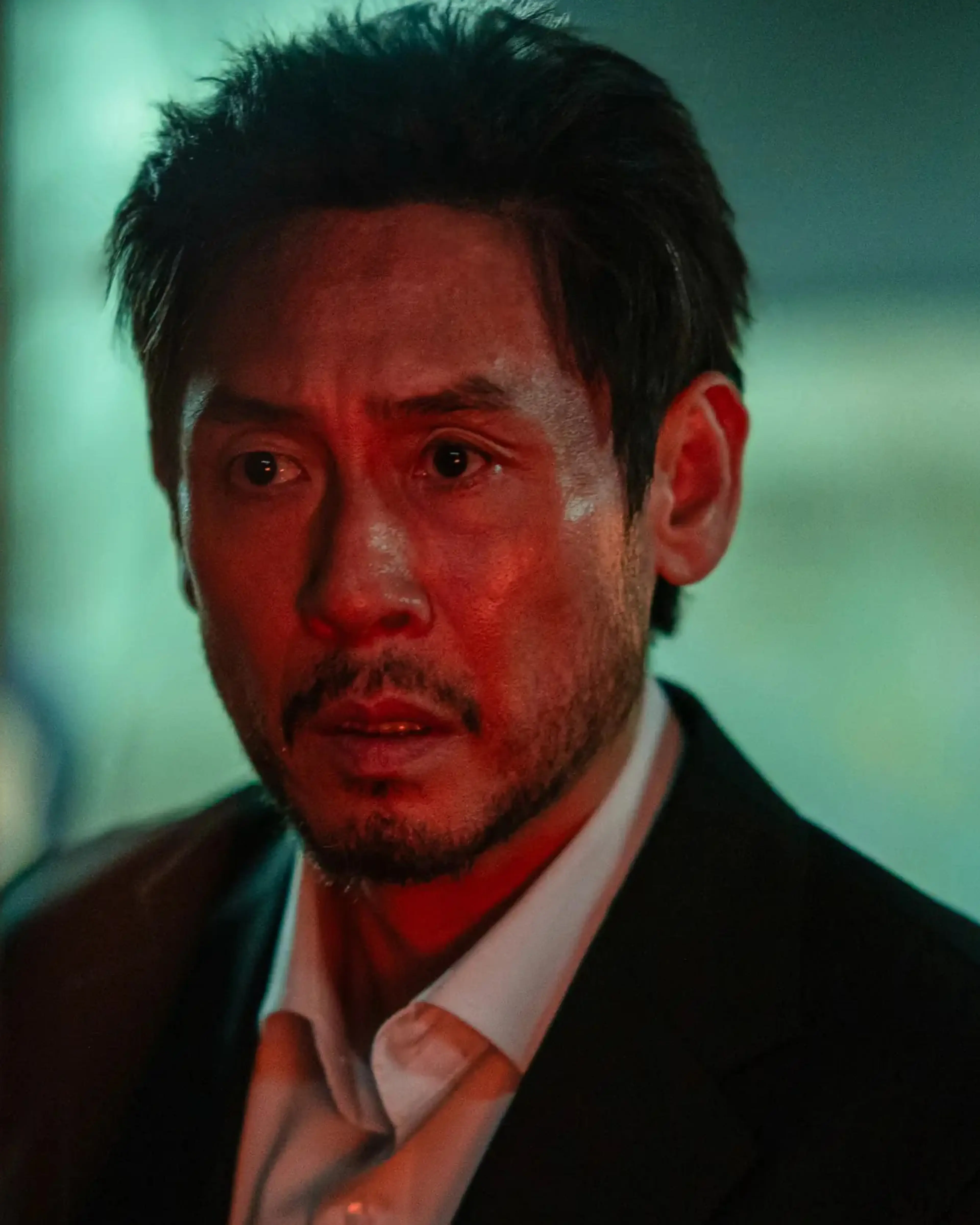 Noja looks ahead while wearing a dark suit and an open-collared white shirt.