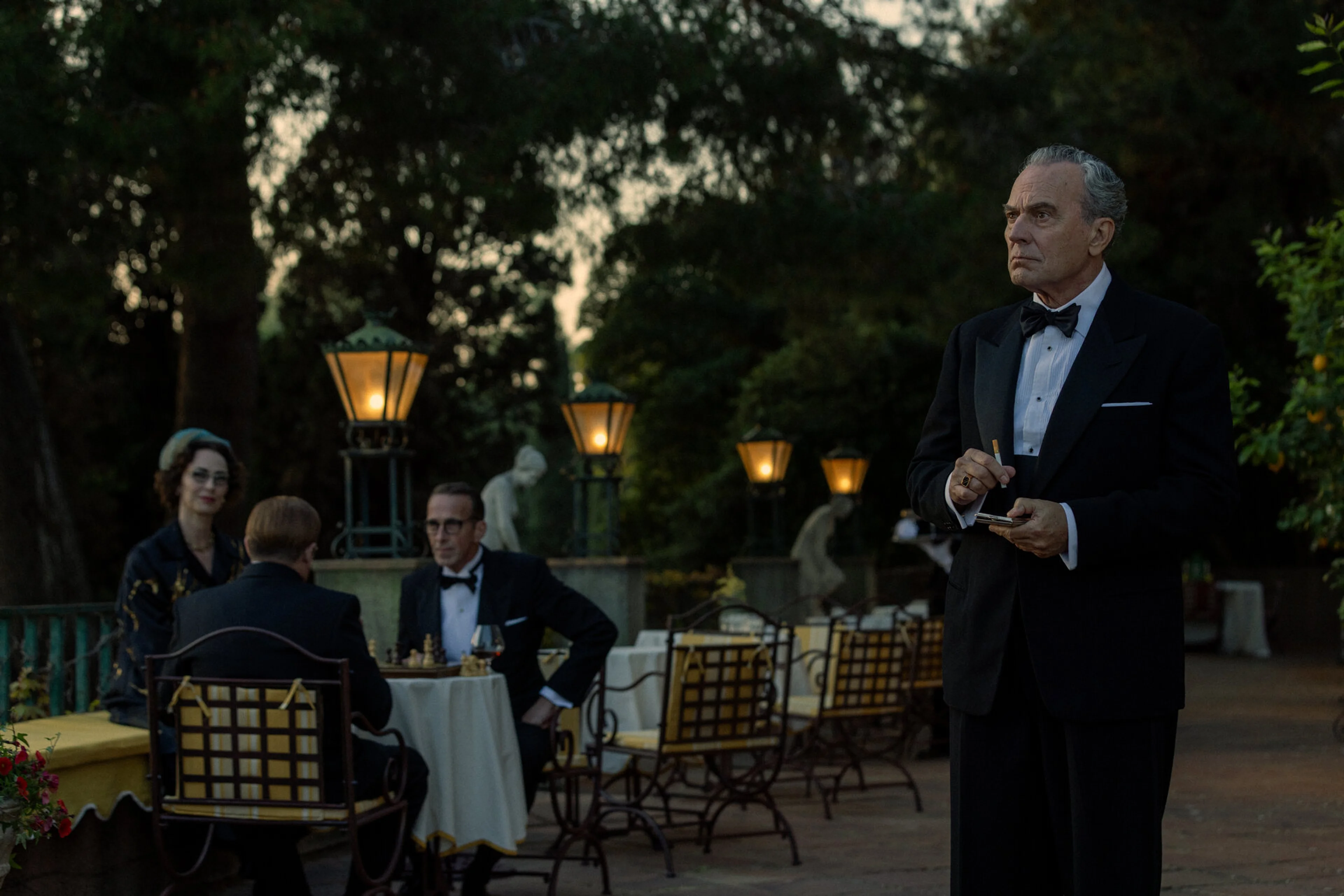 Basil stands on a terrace beside hotel guests in evening clothes.