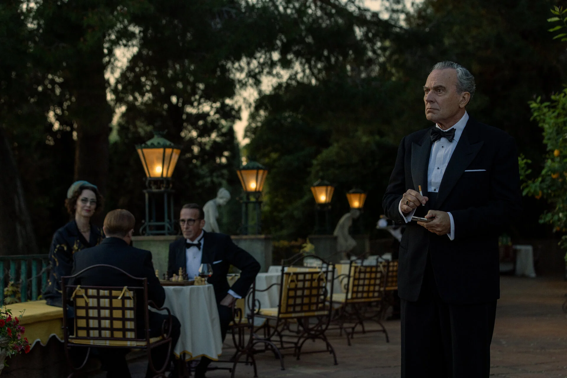 Basil stands on a terrace beside hotel guests in evening clothes.