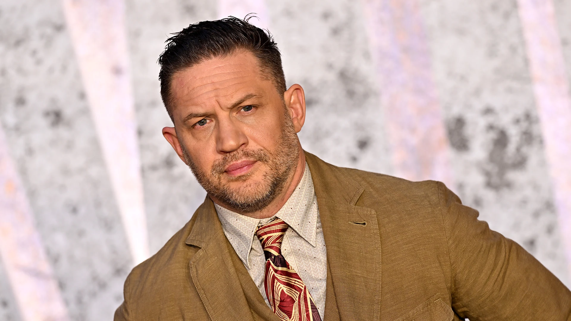 Tom Hardy poses in a tan suit in a portrait used for his Frankie Pulitzer rap album coverage.